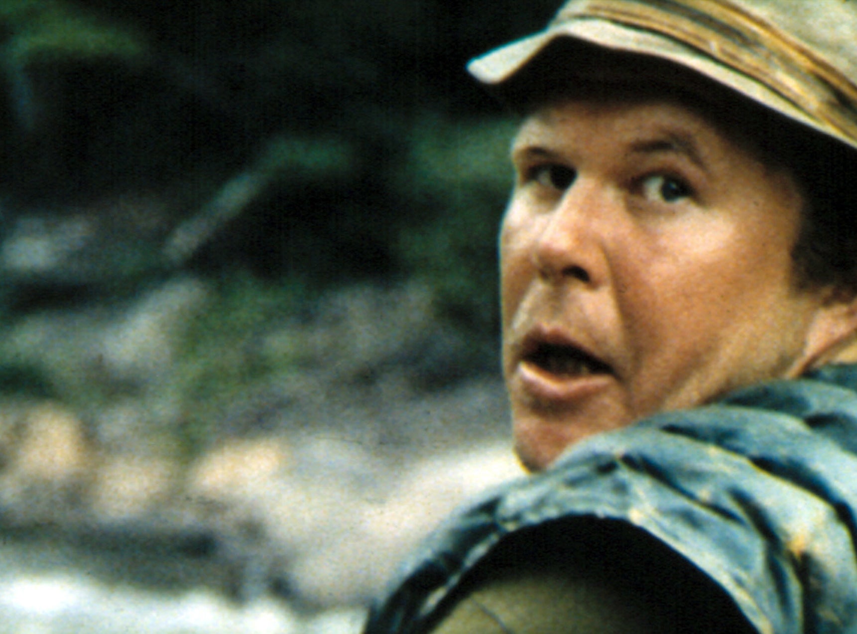 Ned Beatty in an outdoor scene wearing a bucket hat and a quilted jacket, appearing surprised while looking back over his shoulder