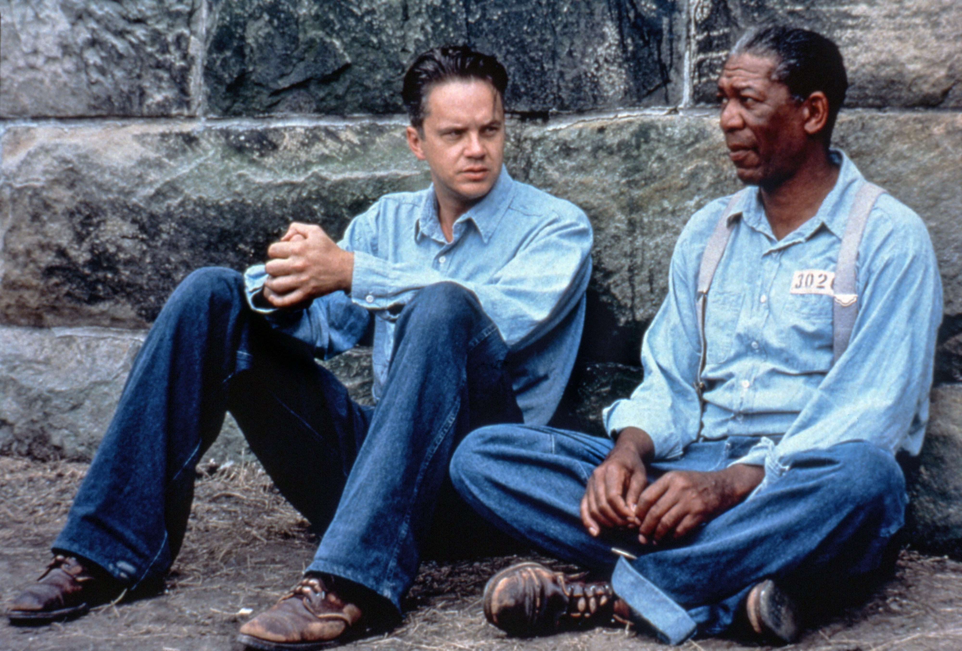 Tim Robbins and Morgan Freeman sit against a stone wall, both wearing prison-style clothes, in a scene from the movie The Shawshank Redemption