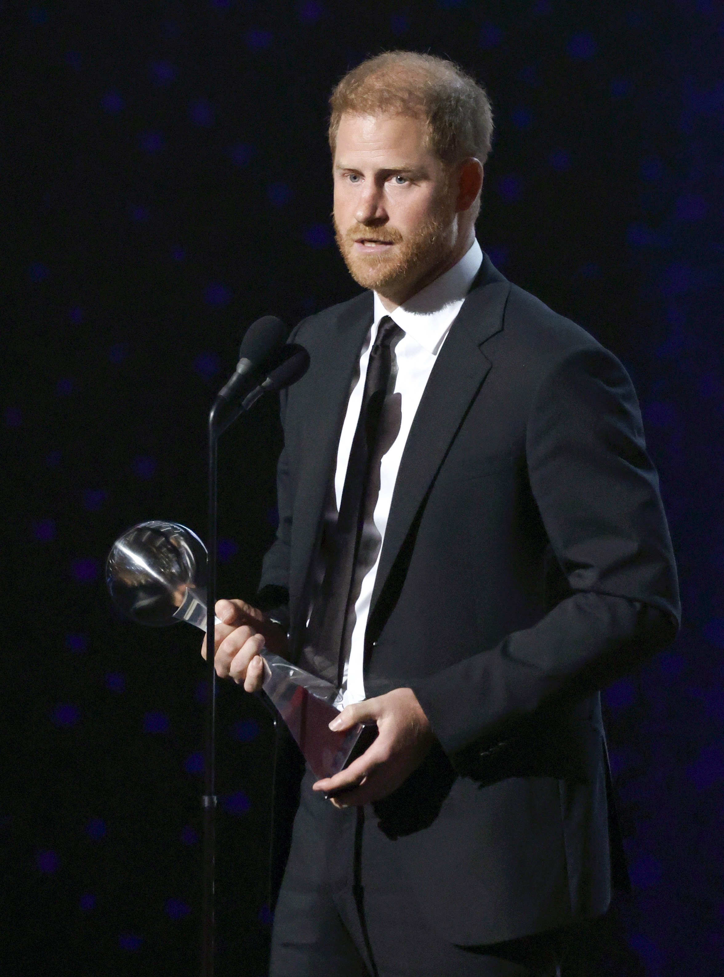Prince Harry in a suit holds a trophy while speaking into a microphone on stage