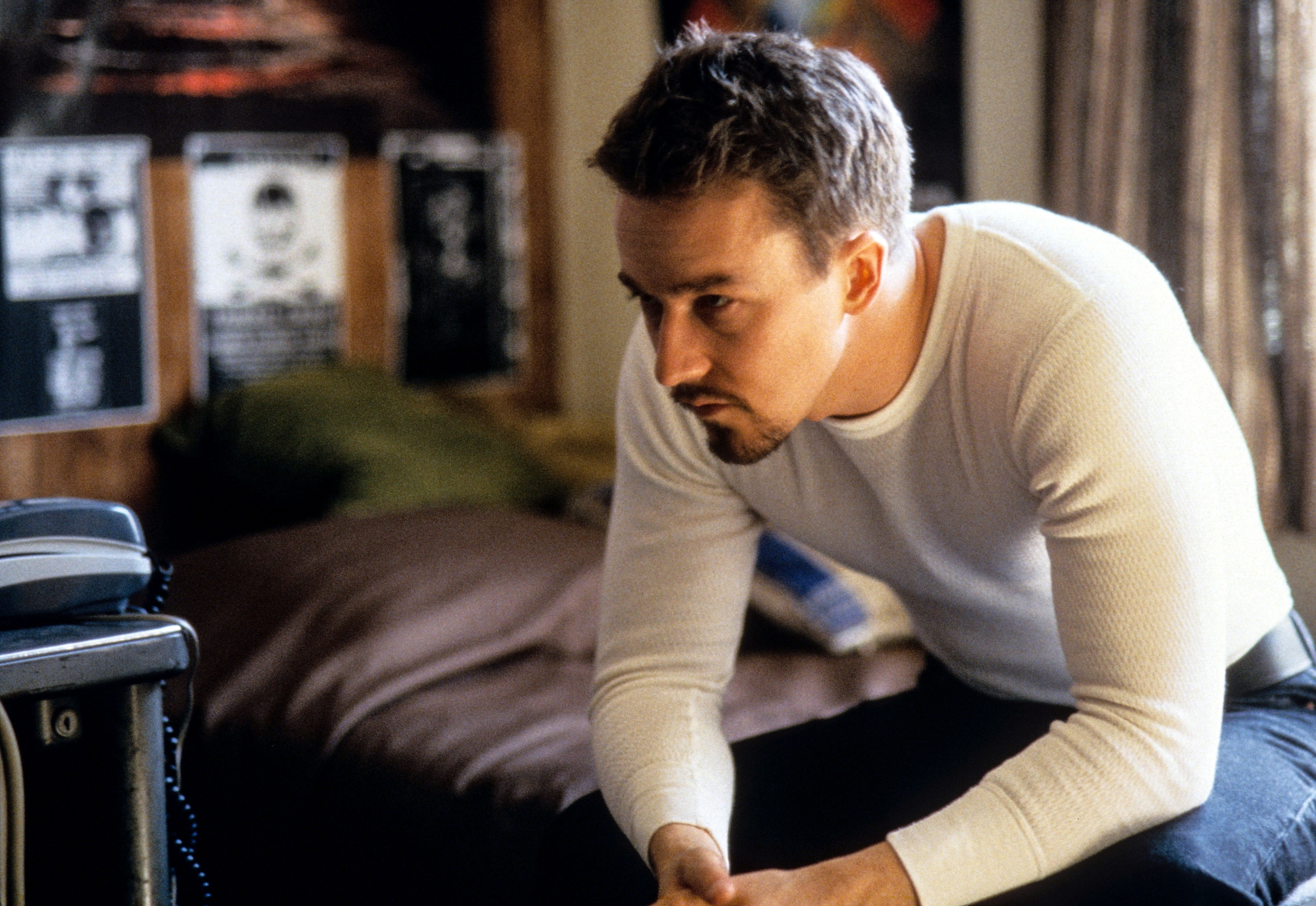 Edward Norton sits on the edge of a bed in a pensive mood, wearing a white long-sleeve shirt. Background features posters on a wall