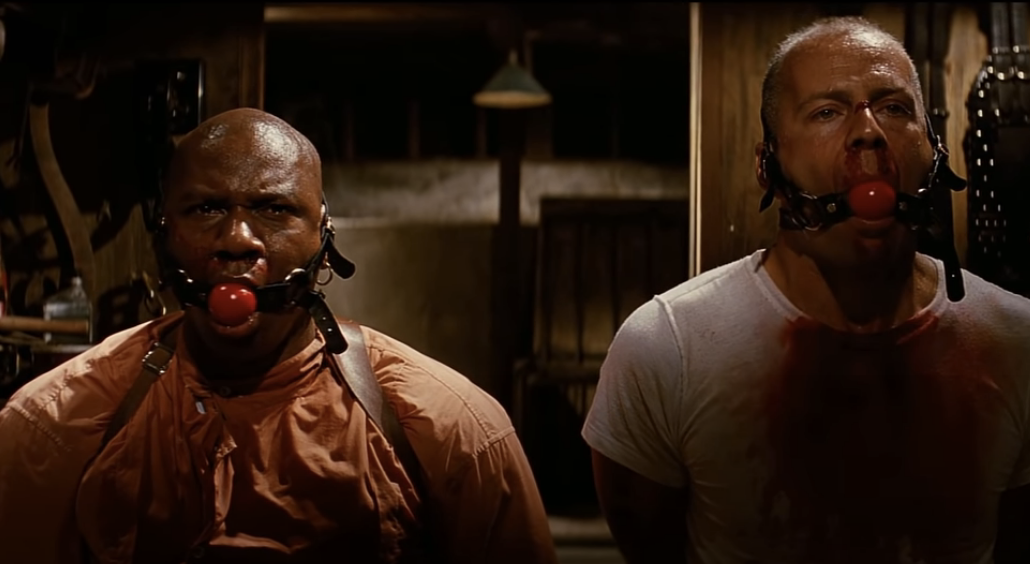 Ving Rhames and Bruce Willis in a scene from a movie, both wearing ball gags in their mouths, standing side by side in a dimly lit room