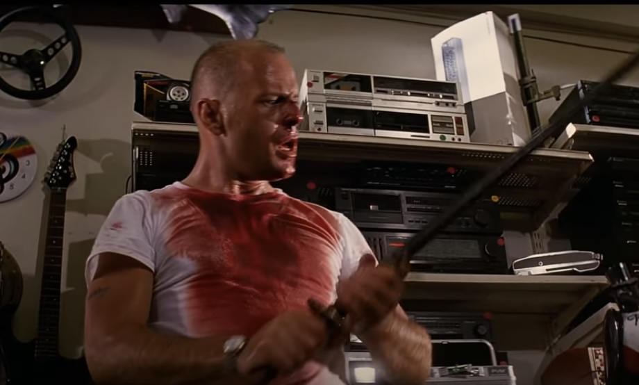 Bruce Willis, in a white t-shirt stained with blood, holds a sword in an intense scene. He is surrounded by electronic equipment, making the setting appear cluttered