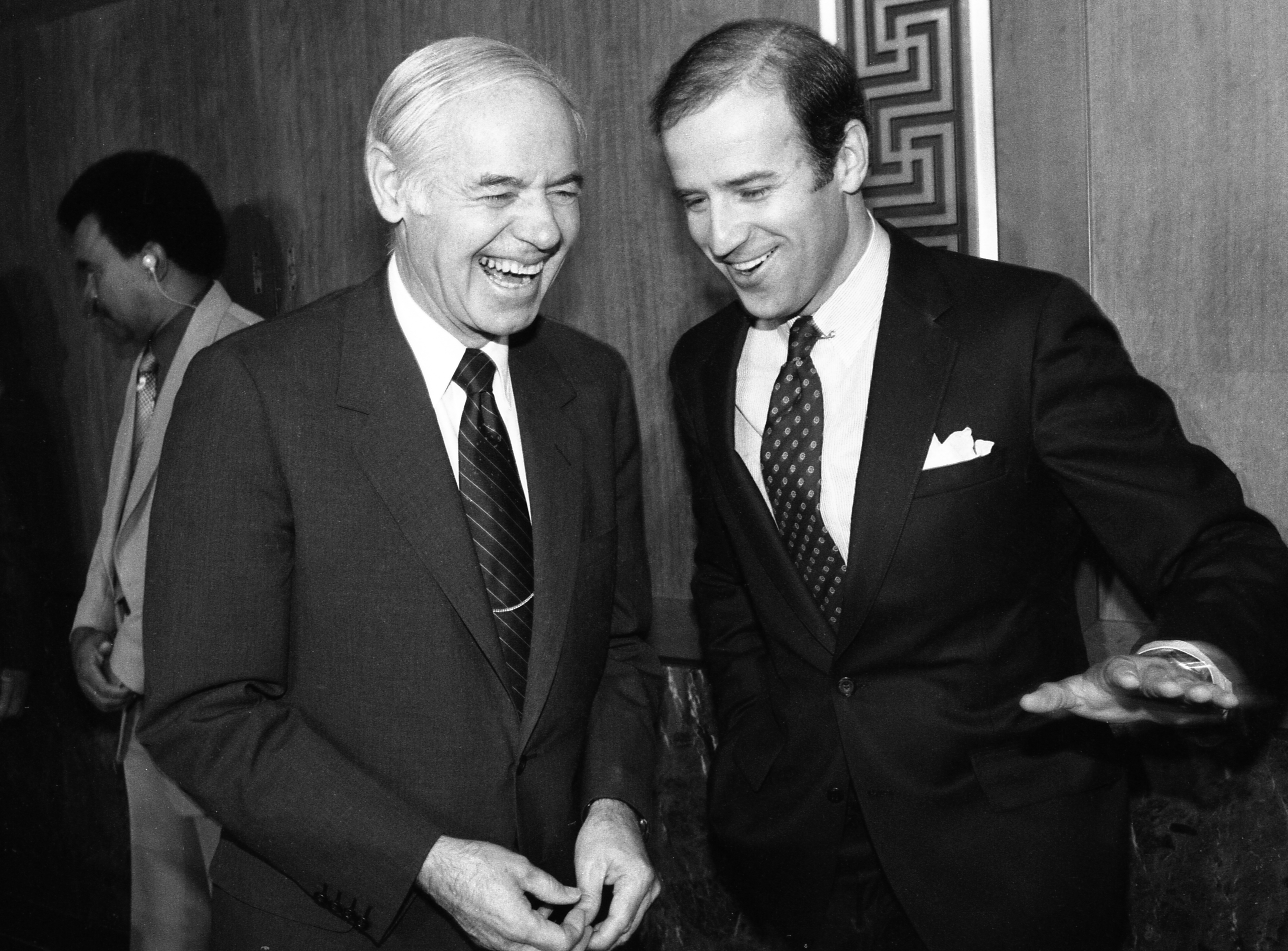 Joe Biden and John Glenn are engaged in conversation, both smiling and wearing suits. A person stands in the background