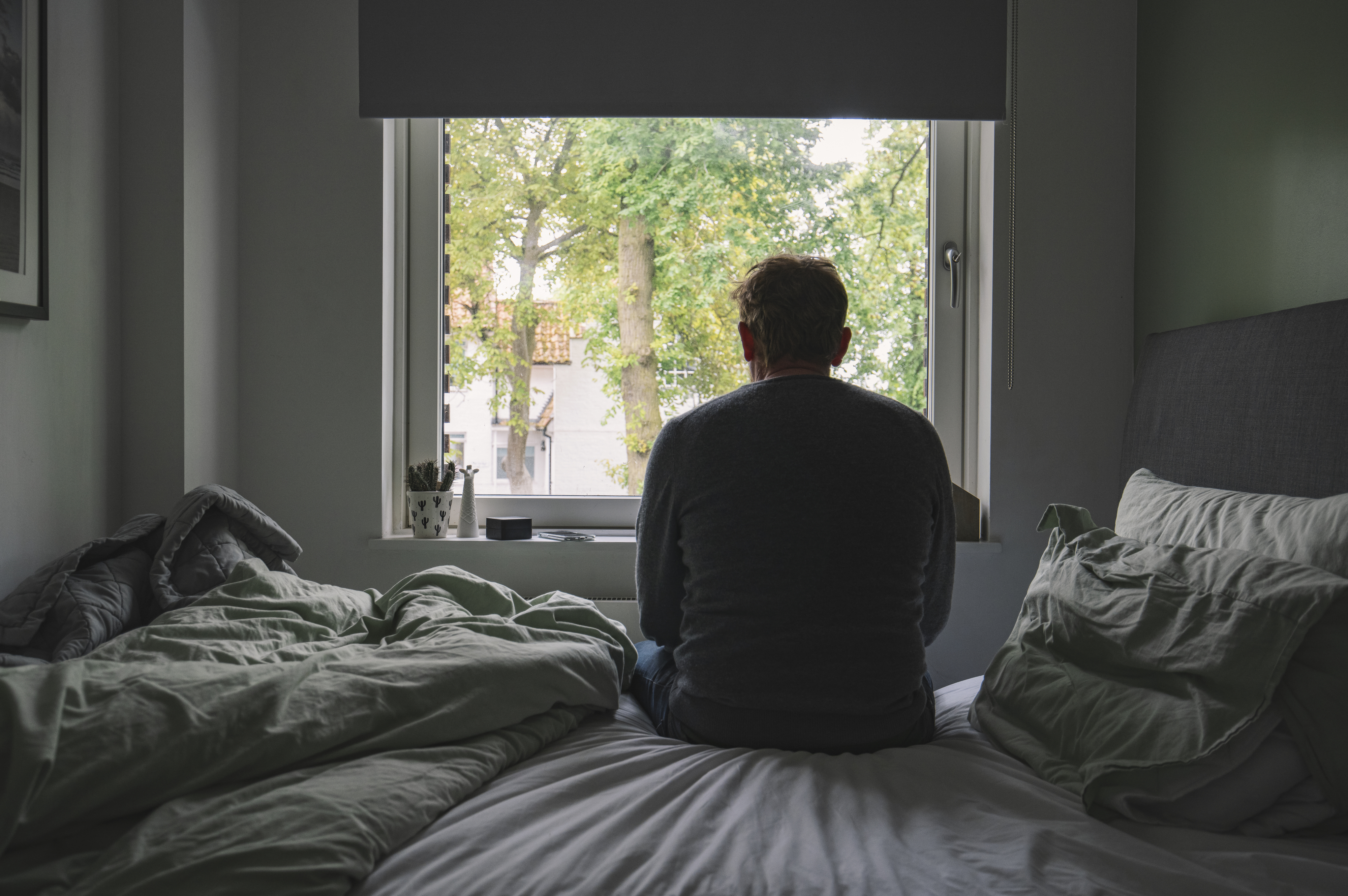 Man sitting on a bed, facing a window with trees outside