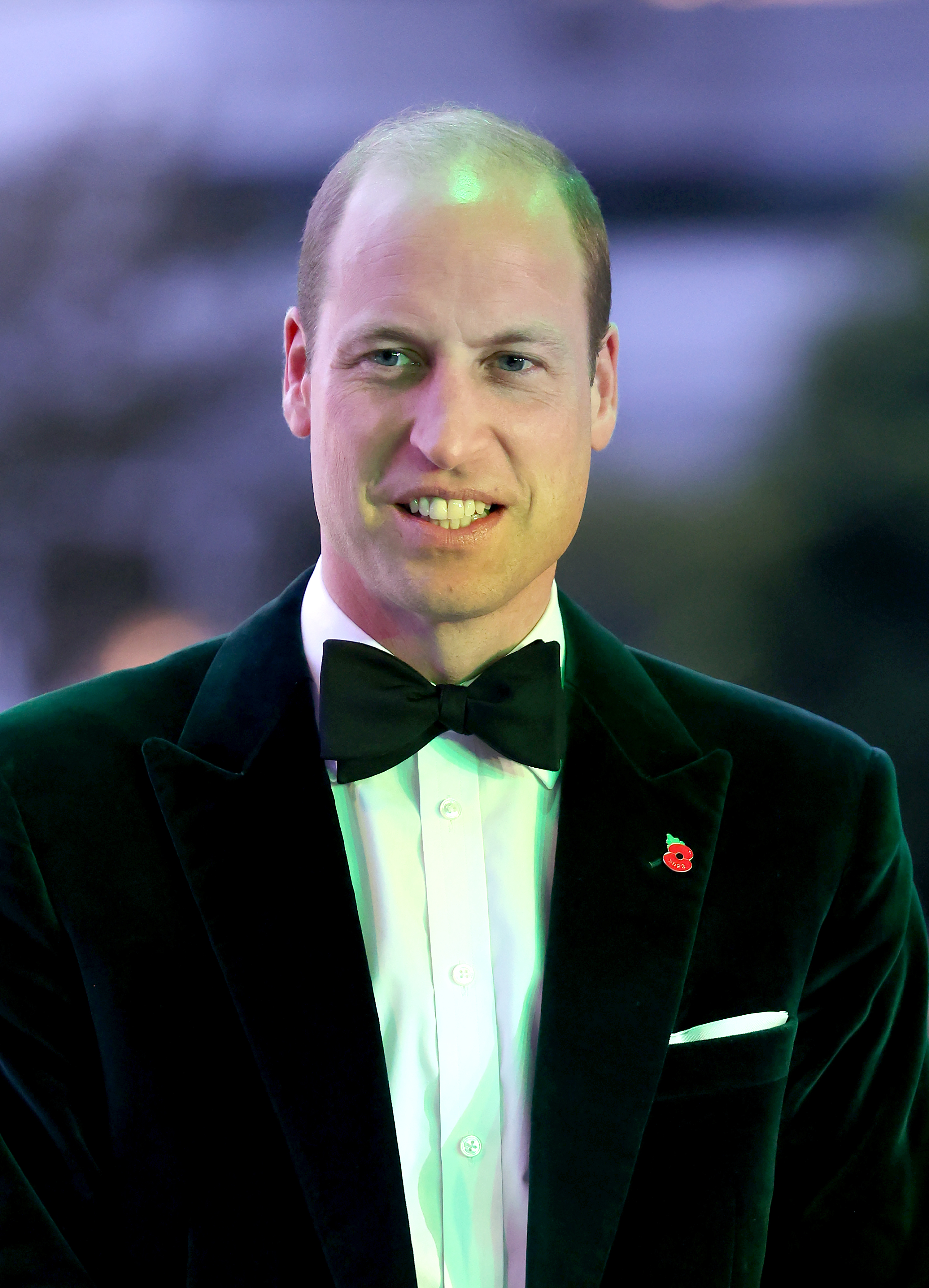 Prince William, in a classic black tuxedo with a bow tie, smiles at the camera on the red carpet