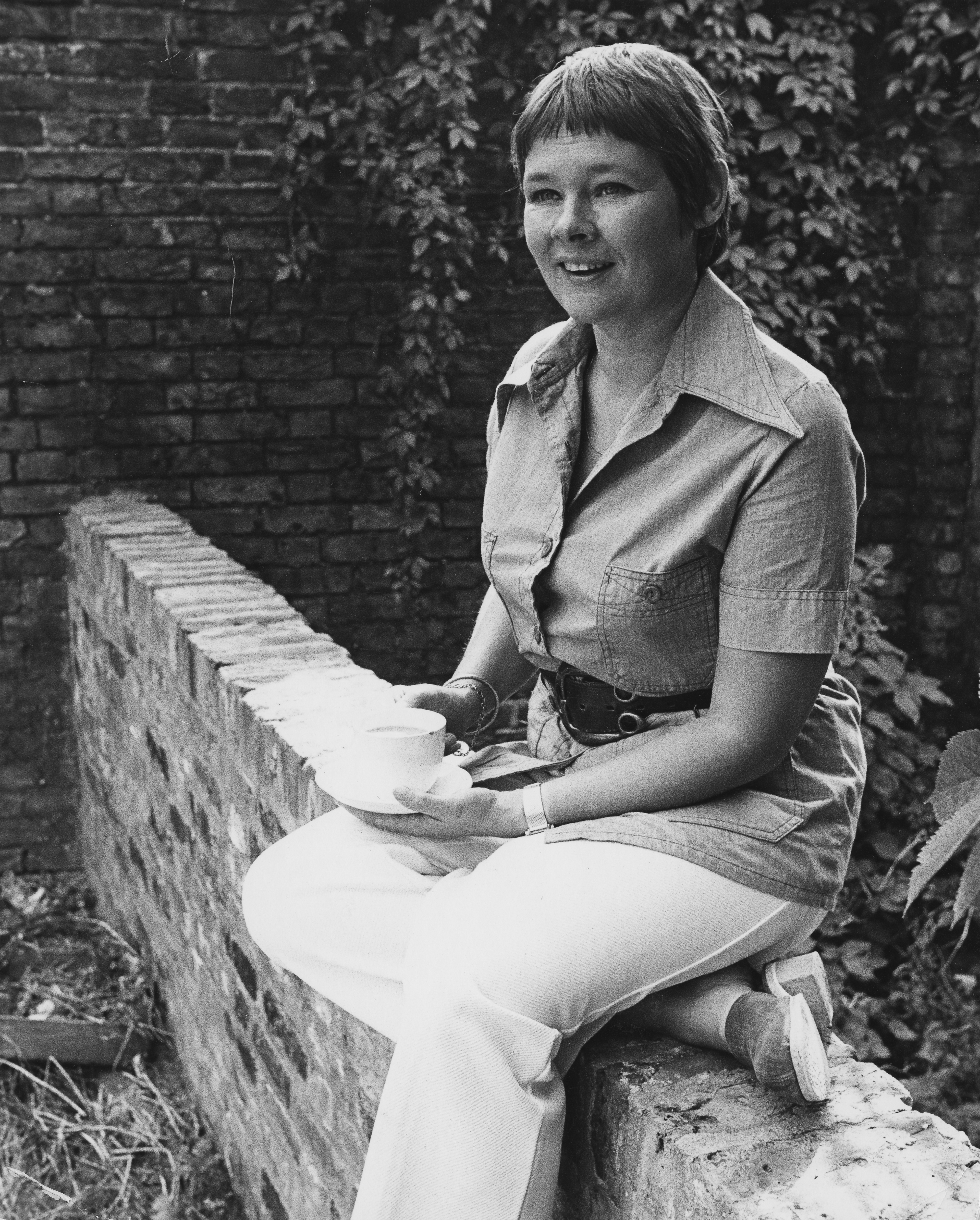 Judi Dench sits on a brick wall, holding a teacup, in a belted shirt and light-colored pants, with ivy-covered walls in the background
