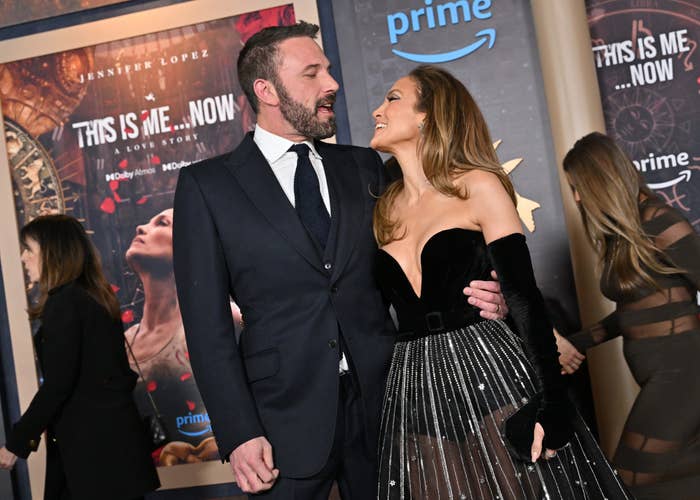 Ben Affleck in a dark suit and Jennifer Lopez in a gown with a sheer skirt smile at each other at the "This Is Me…Now" event