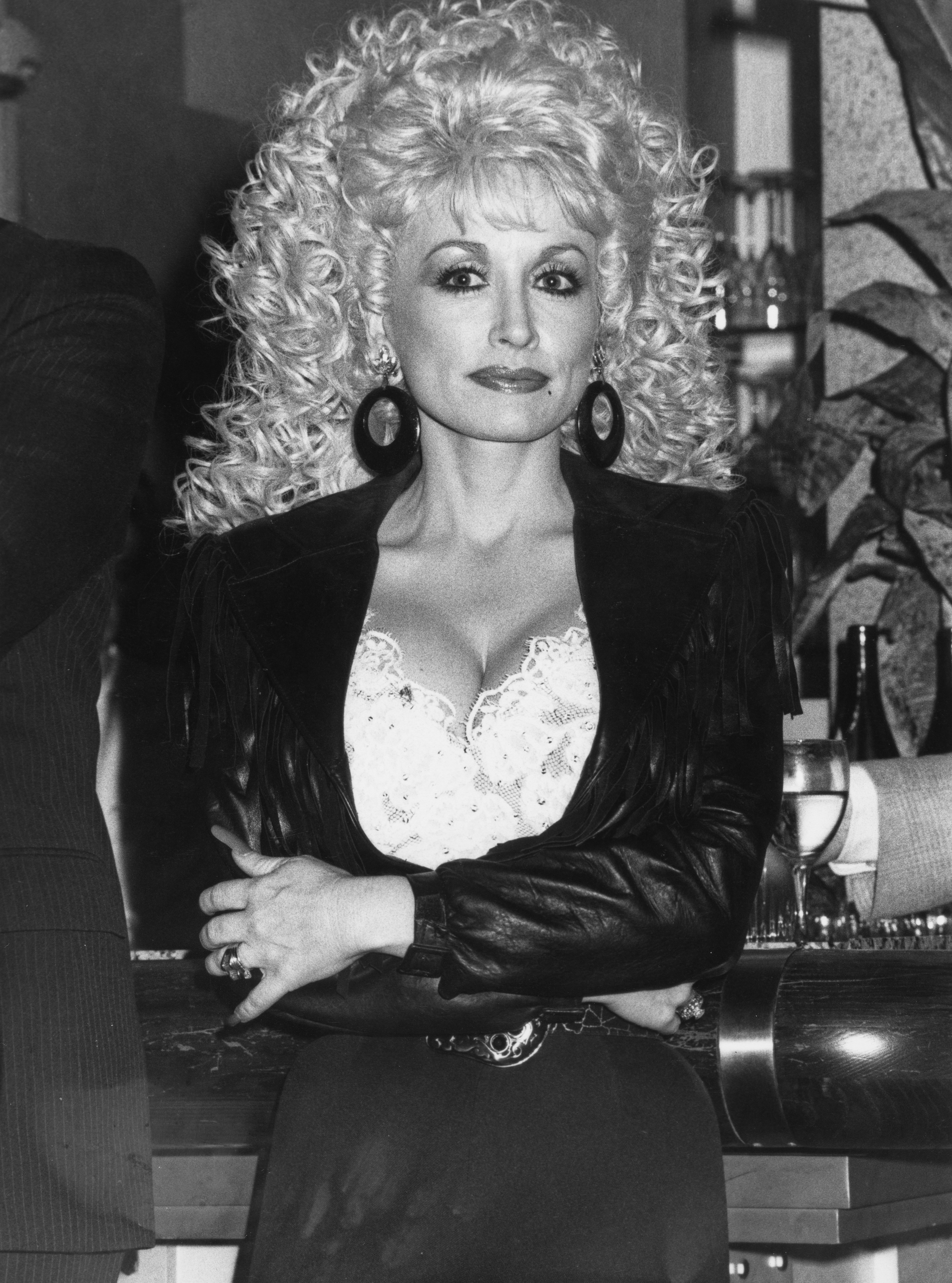 Dolly Parton stands with arms crossed, wearing a fringed jacket and lace top, with large earrings and voluminous curly hair in a classic, stylish look