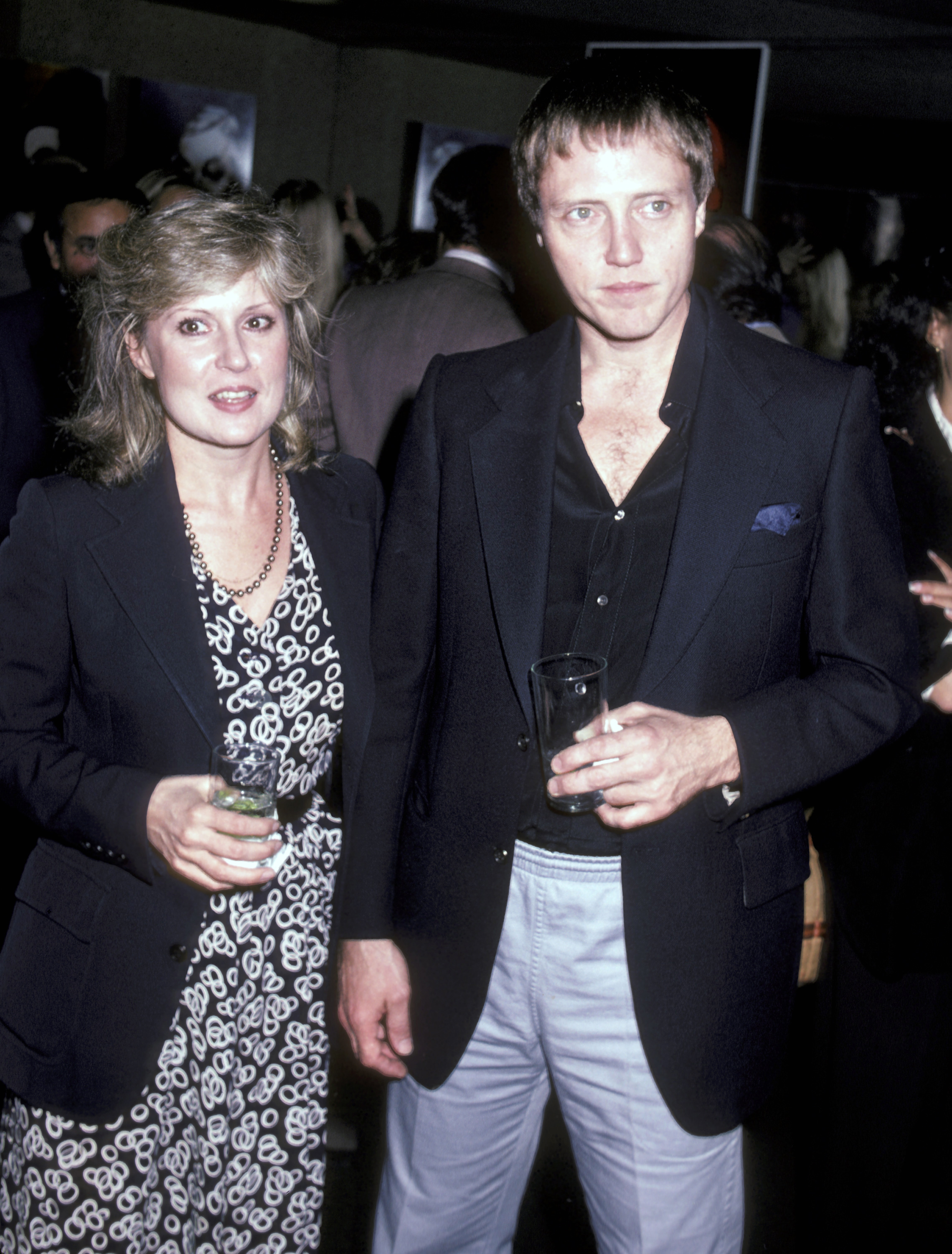 Nancy Allen and Christopher Walken at an event, Nancy wearing a patterned dress and dark blazer, Christopher in a dark blazer and light pants, both holding drinks