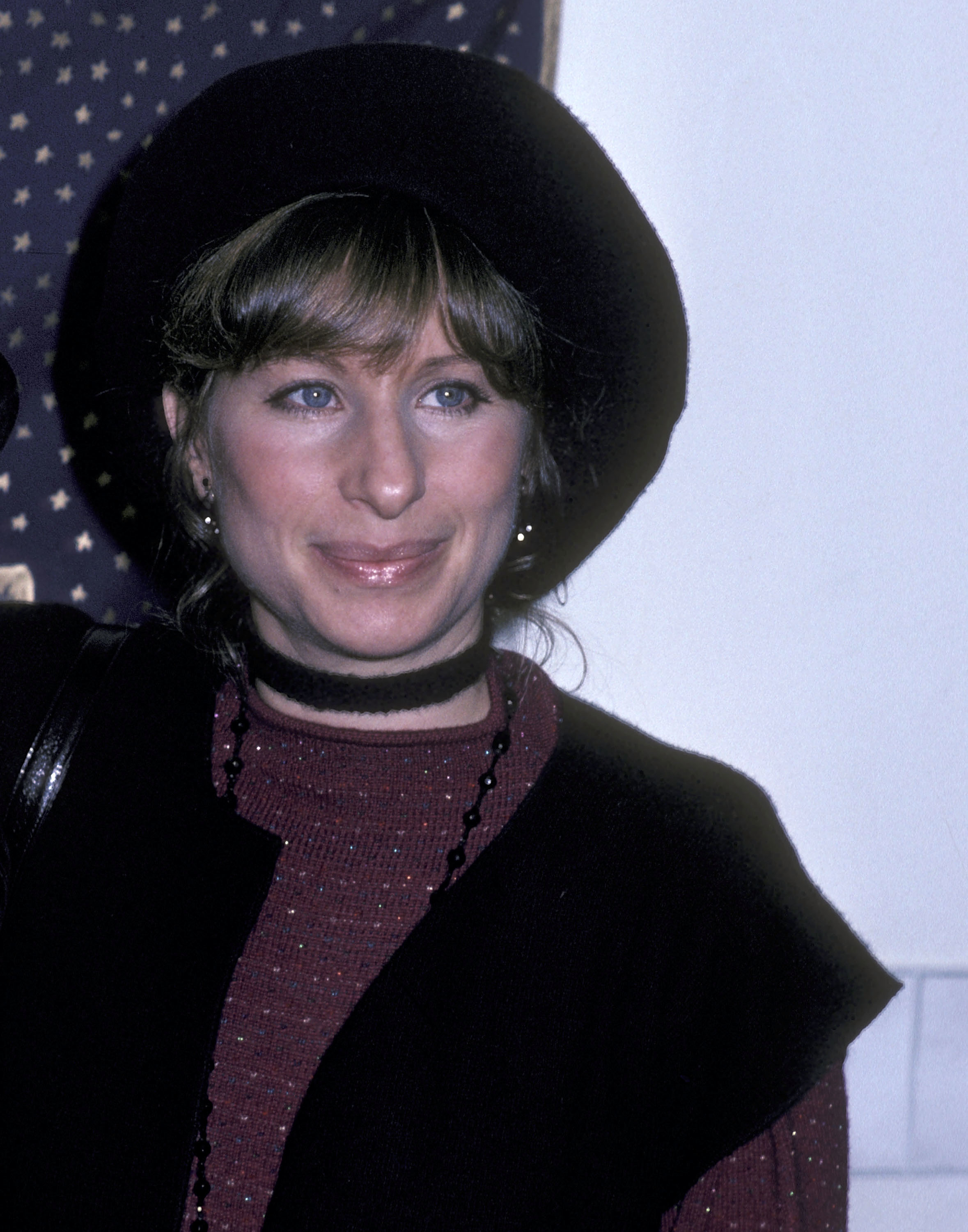 Barbra Streisand is seen wearing a hat, a round-neck top, and a black vest with a necklace in this image