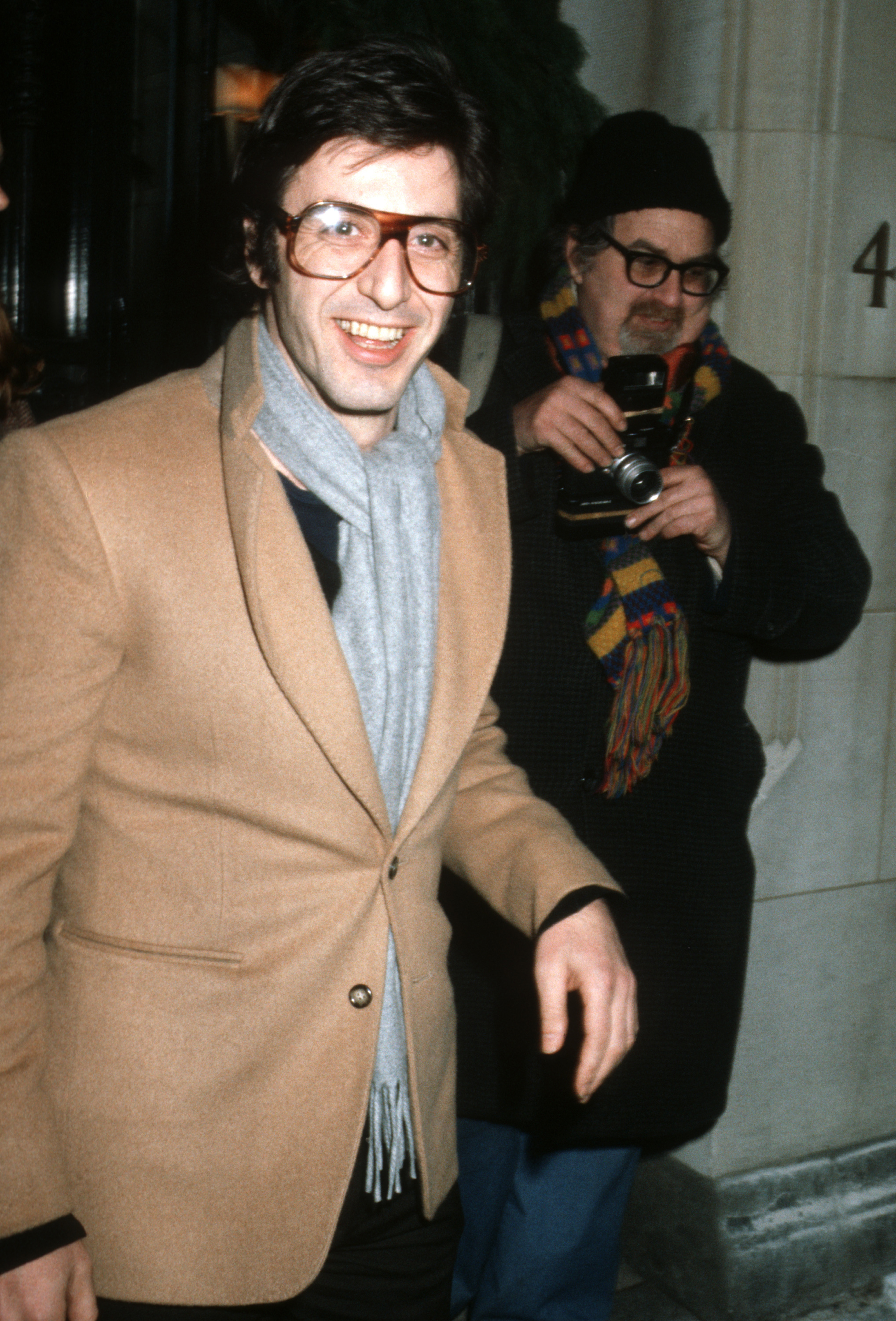 Al Pacino smiles while wearing a tailored jacket and scarf. Another person behind him holds a camera, wearing a hat and colorful scarf