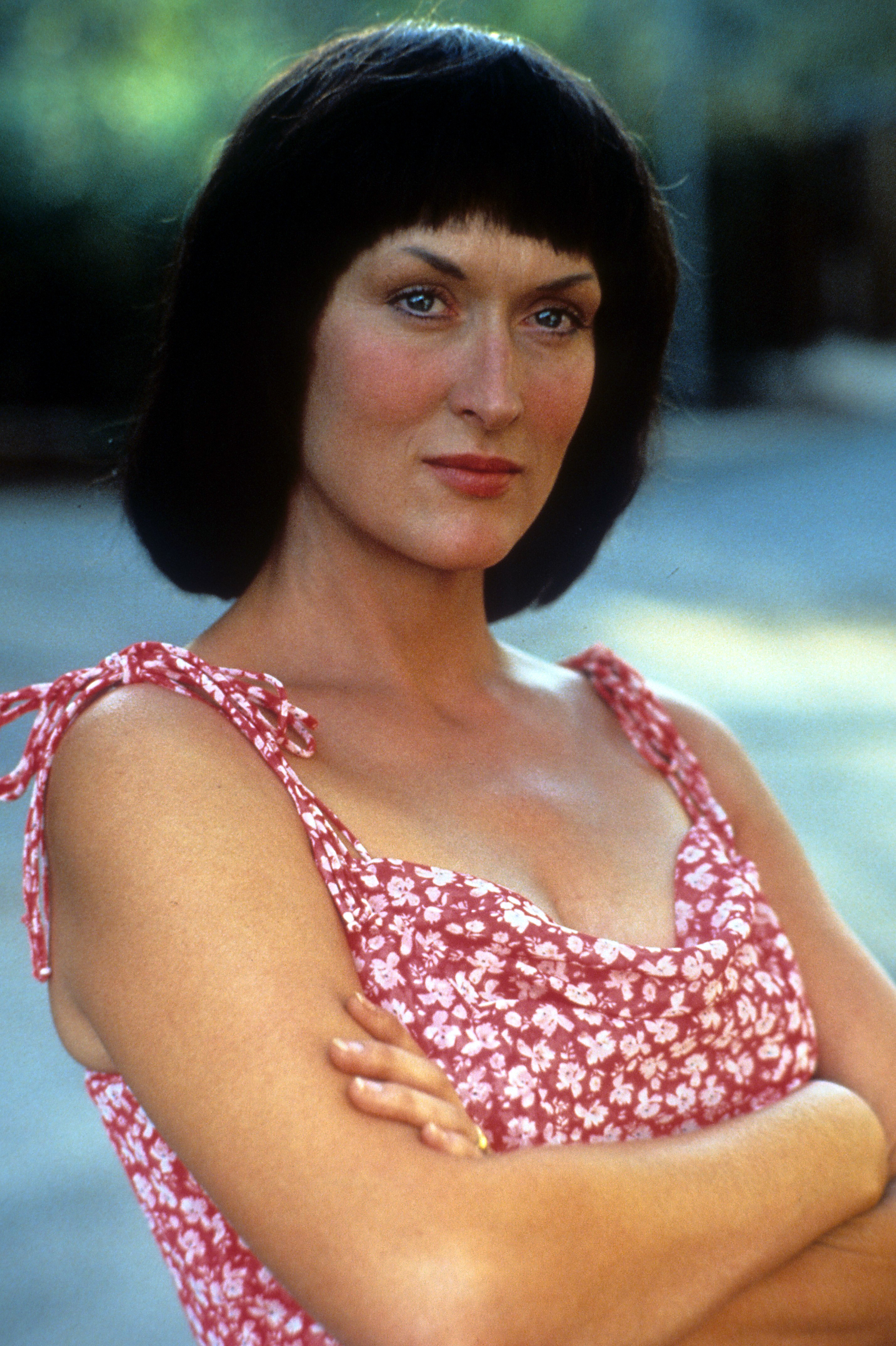 Meryl Streep with short dark hair and bangs, wearing a sleeveless floral dress, arms crossed, and looking confidently into the camera