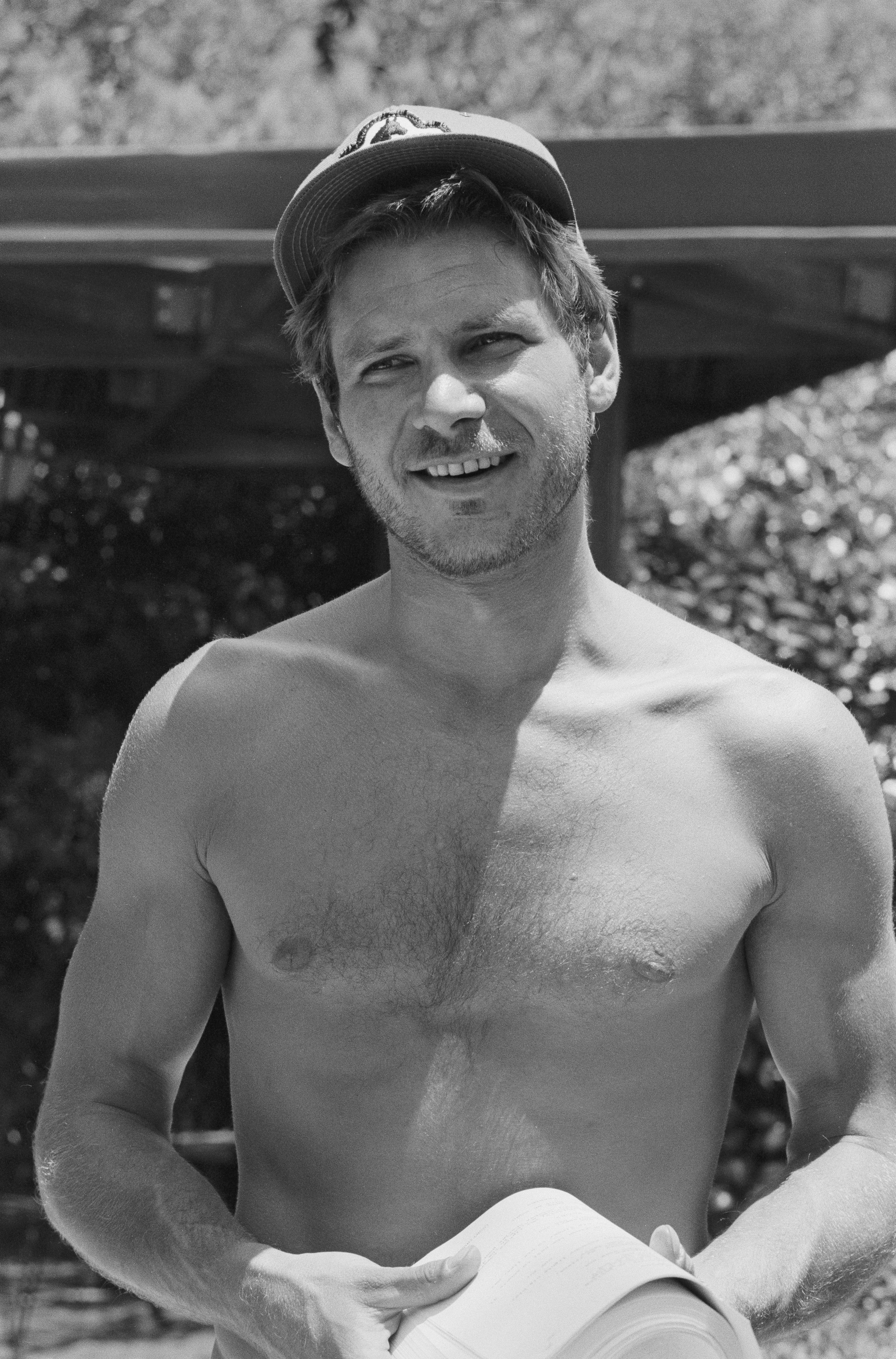 A shirtless Harrison Ford wearing a cap, outdoors, holding a book or paper