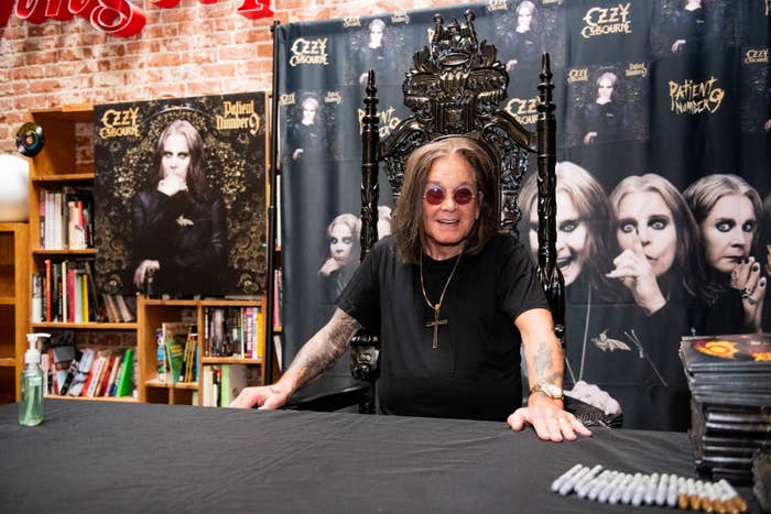 Ozzy Osbourne, wearing a dark t-shirt and gold cross necklace, sits at a table during an autograph signing event in front of his concert posters
