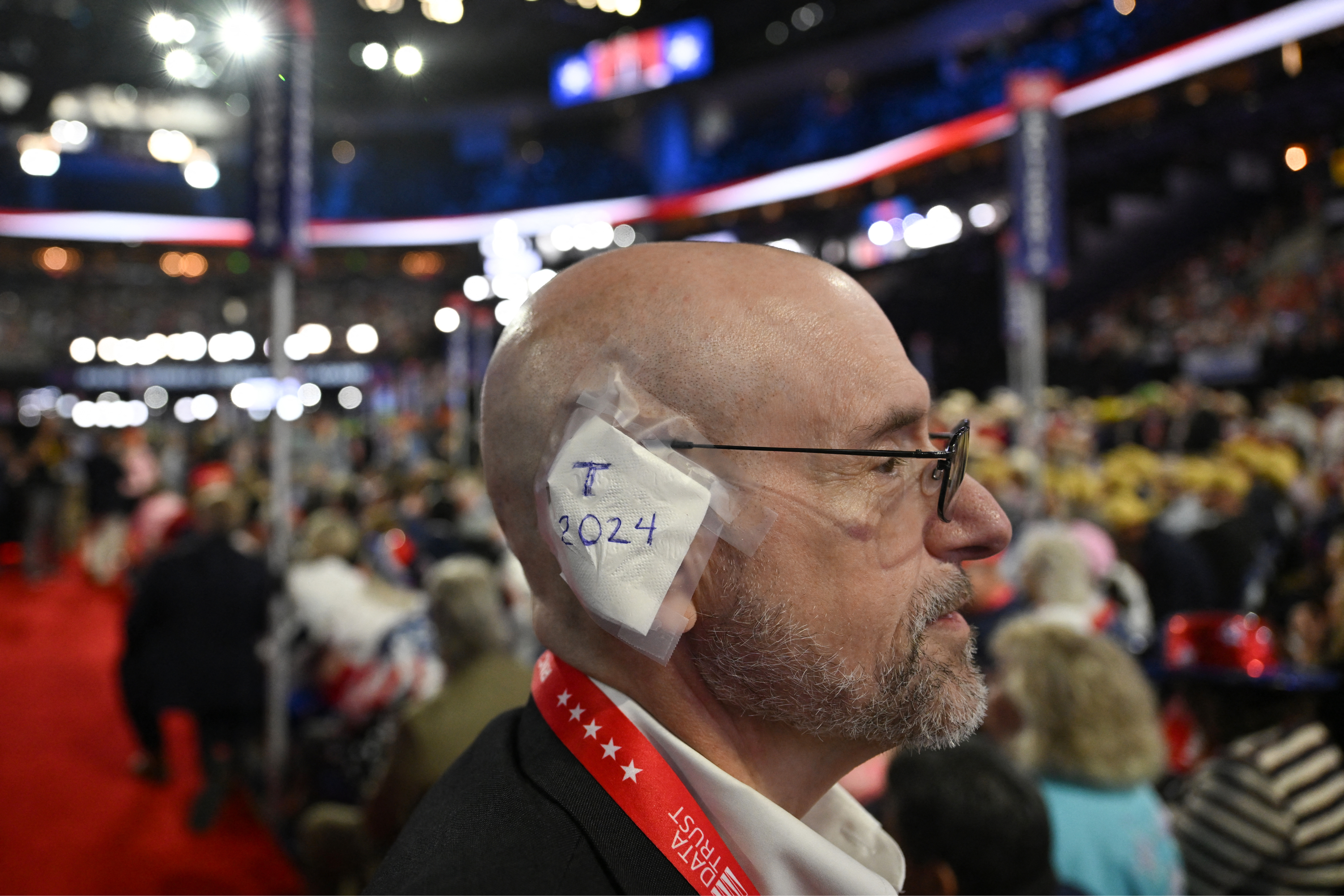 Man with a bald head and glasses at a crowded indoor event, with a sign reading &quot;T 2024&quot; taped to the side of his head