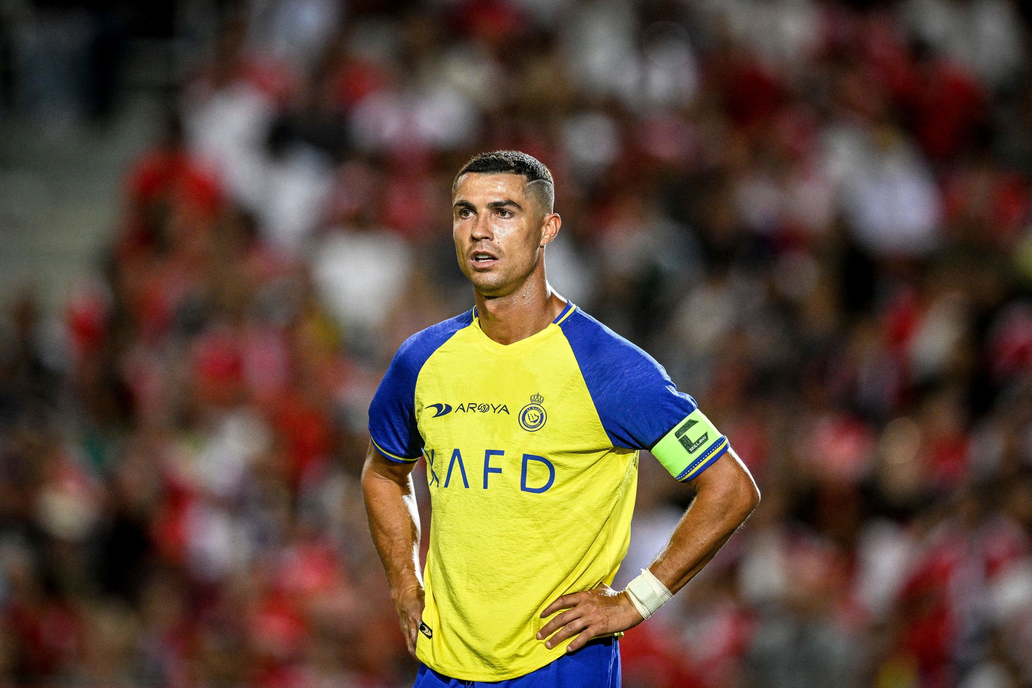 Cristiano Ronaldo stands on a soccer field, wearing an Al-Nassr jersey, with hands on hips and a focused expression