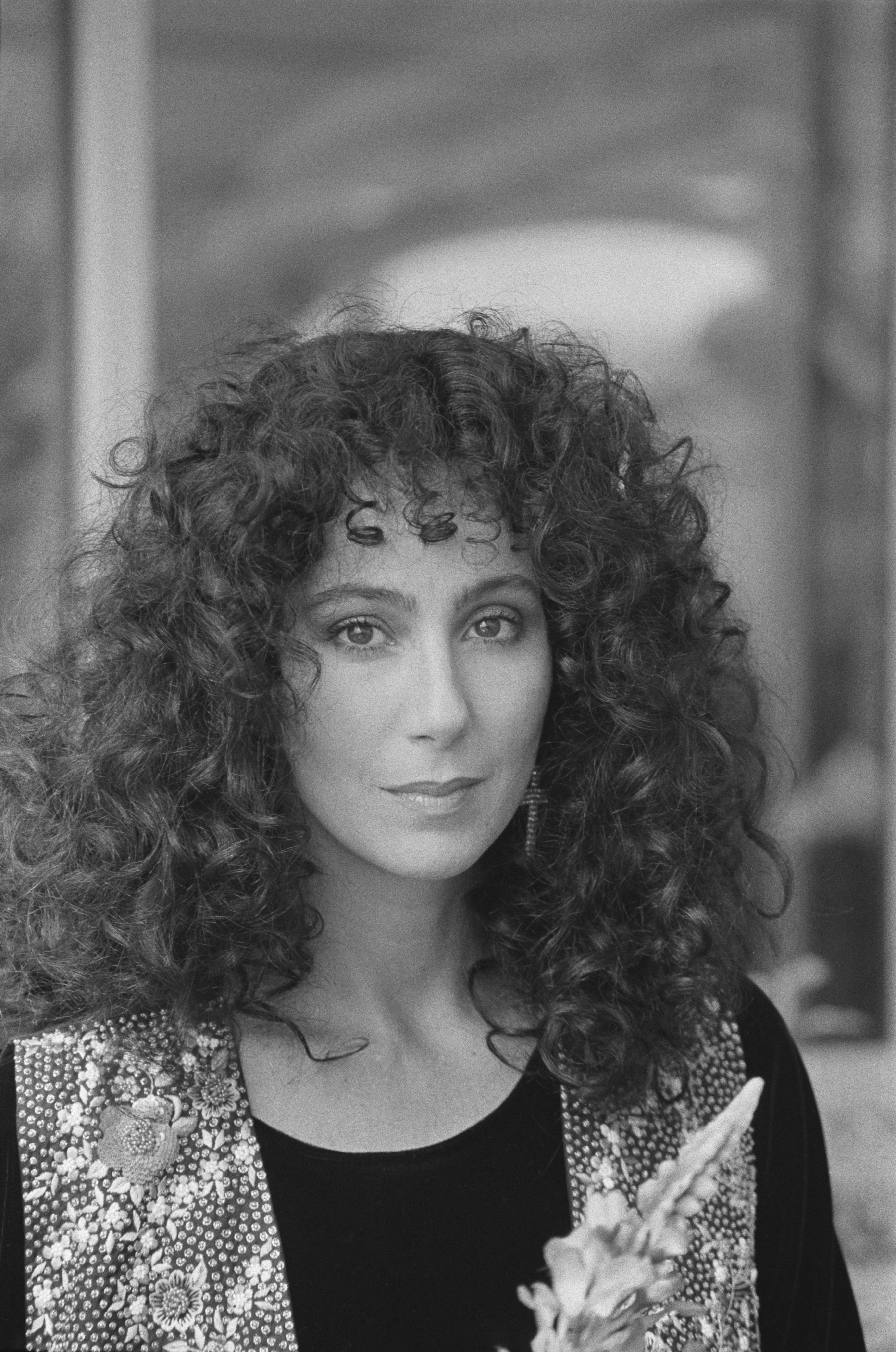 Cher with curly hair, wearing a floral-patterned vest over a black top, gazing directly at the camera