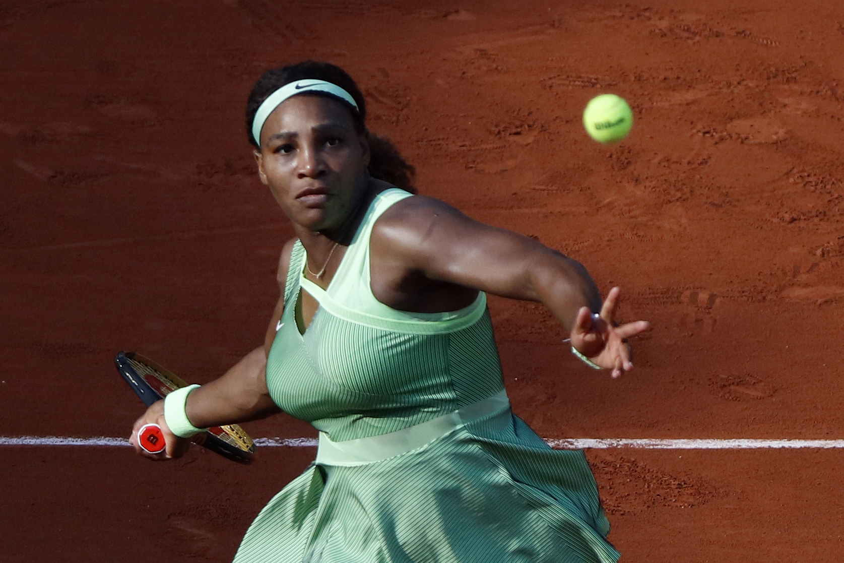 Serena Williams is playing tennis on a clay court, wearing a sleeveless, pleated dress and a headband. She is focused on hitting the incoming tennis ball