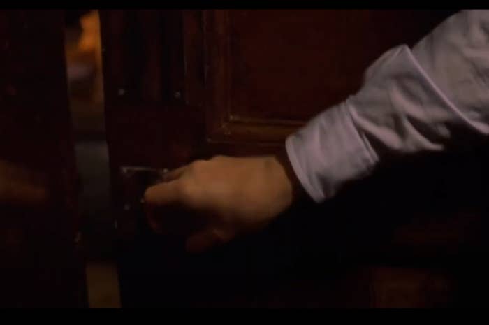 A hand, wearing a white sleeve, partially opens a wooden door by turning the handle
