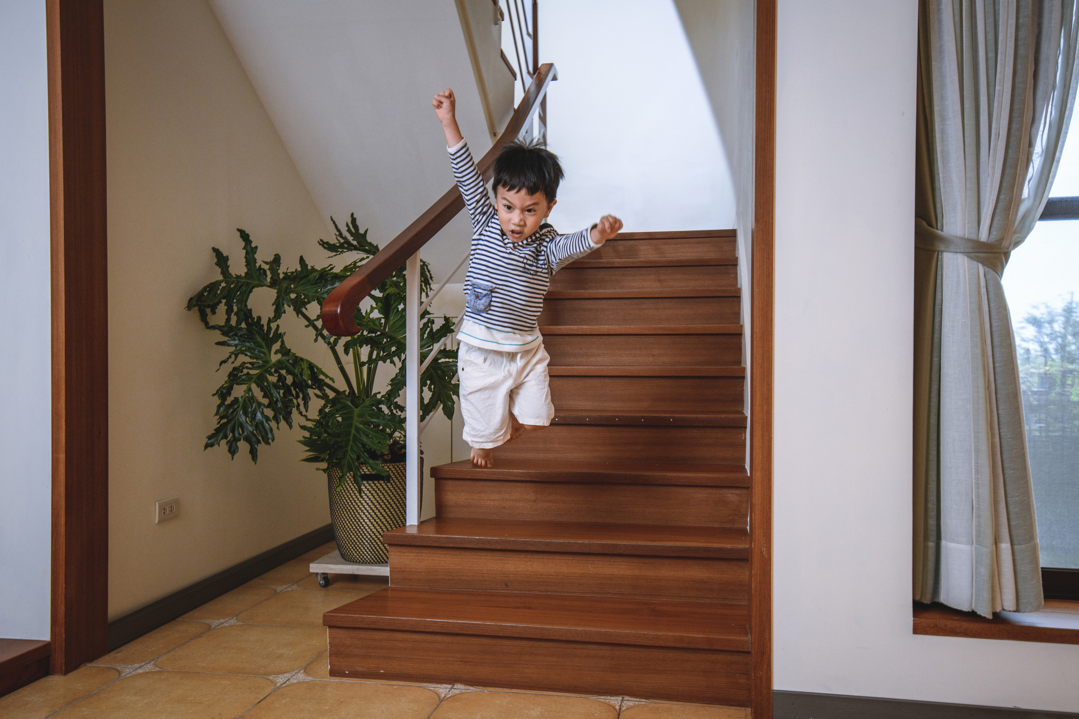 A young child wearing a striped shirt and white shorts joyfully jumps from the stairs inside a home with a potted plant and window in the background