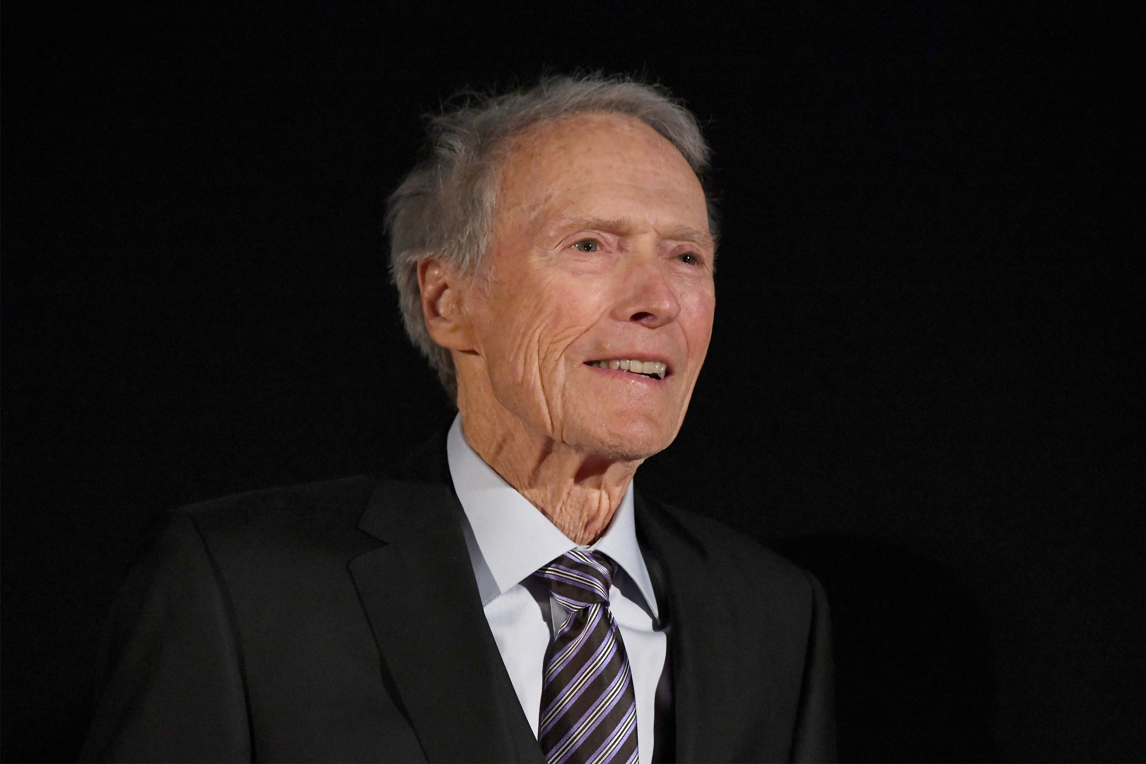 Clint Eastwood wearing a dark suit and striped tie, smiling and looking off-camera against a black background