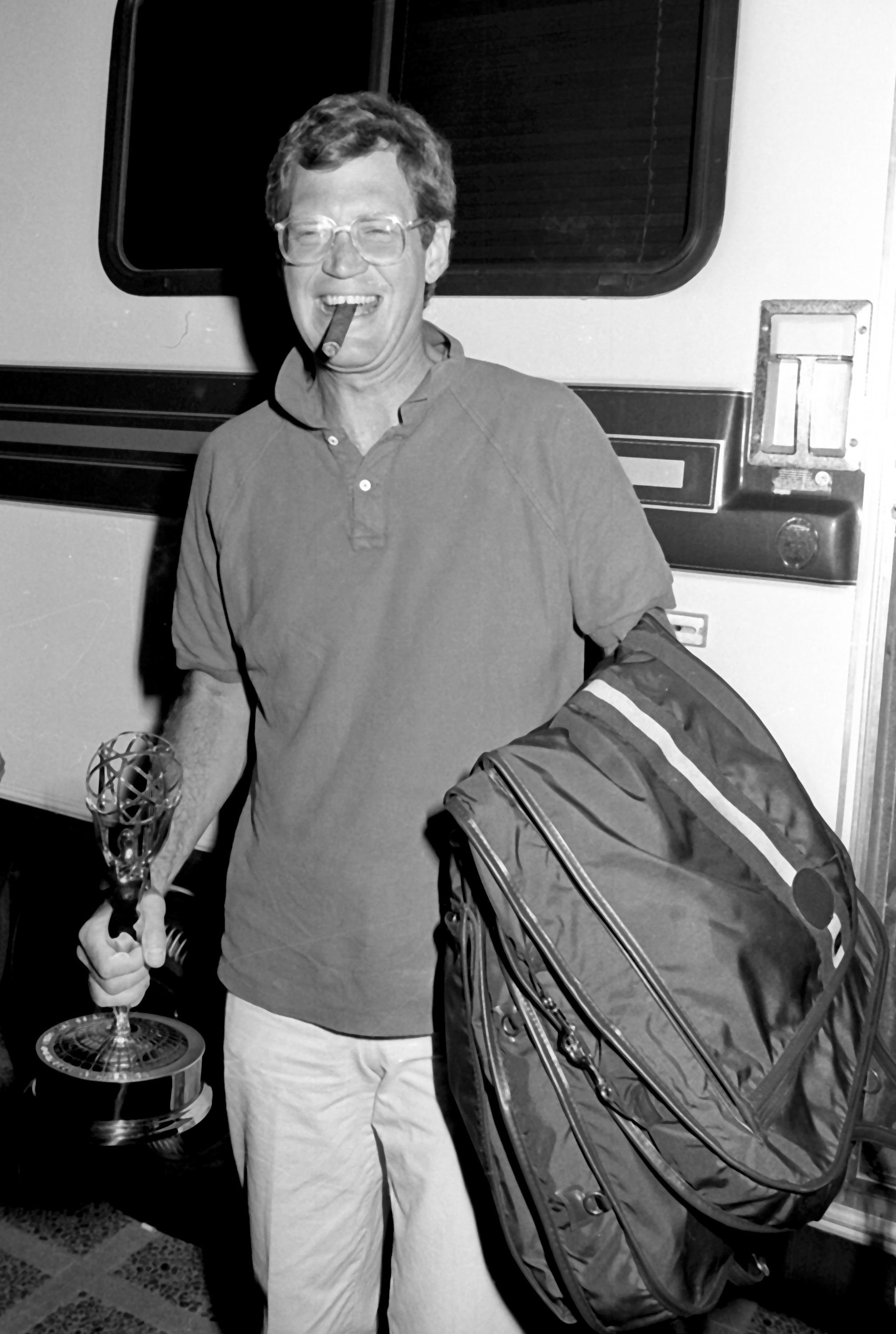A person, wearing a polo shirt and holding an Emmy award in one hand and a bag in the other, smiles widely with a cigar in their mouth