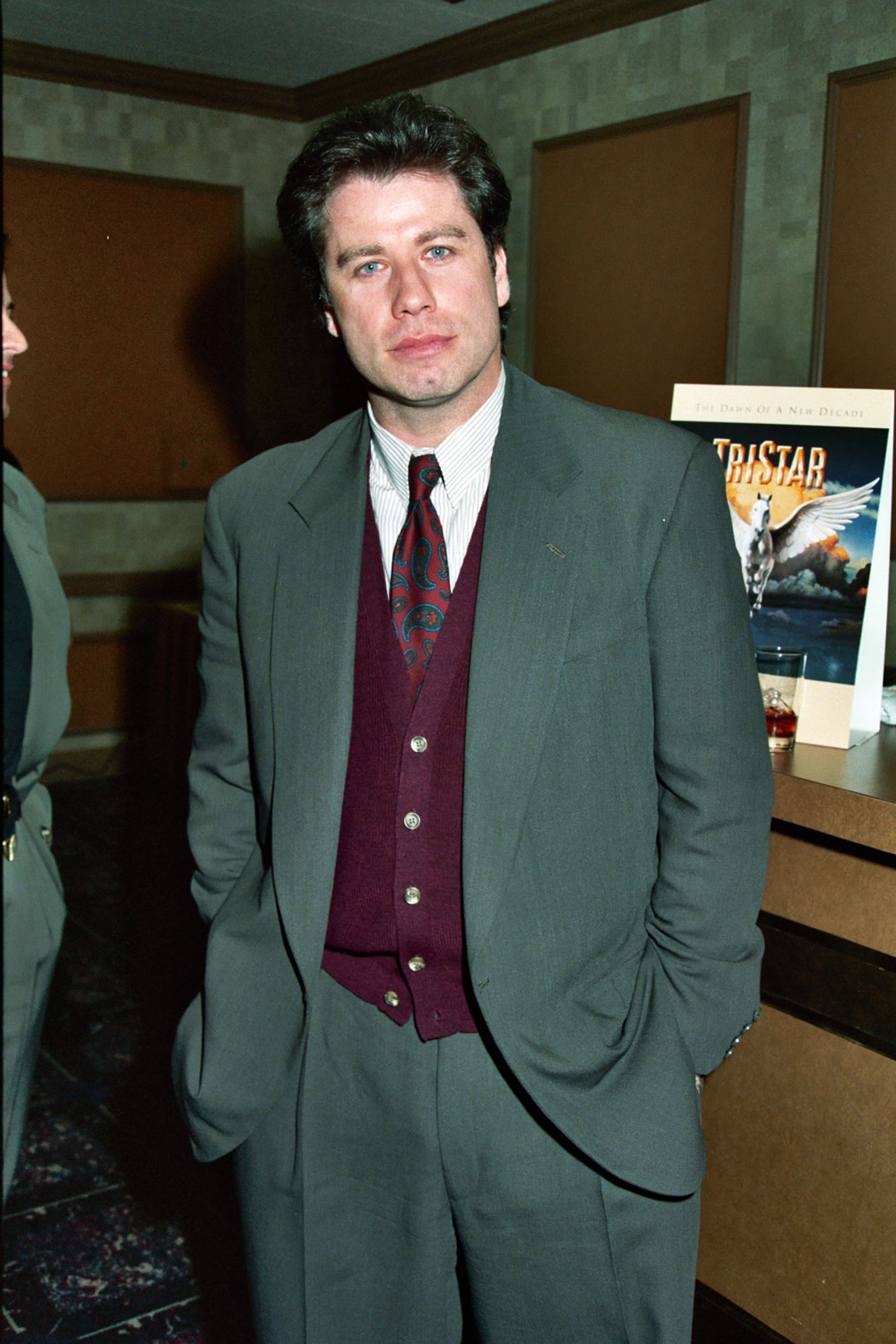 John Travolta in a suit and patterned tie with a burgundy sweater vest, hands in pockets, standing indoors near a Tristar movie poster