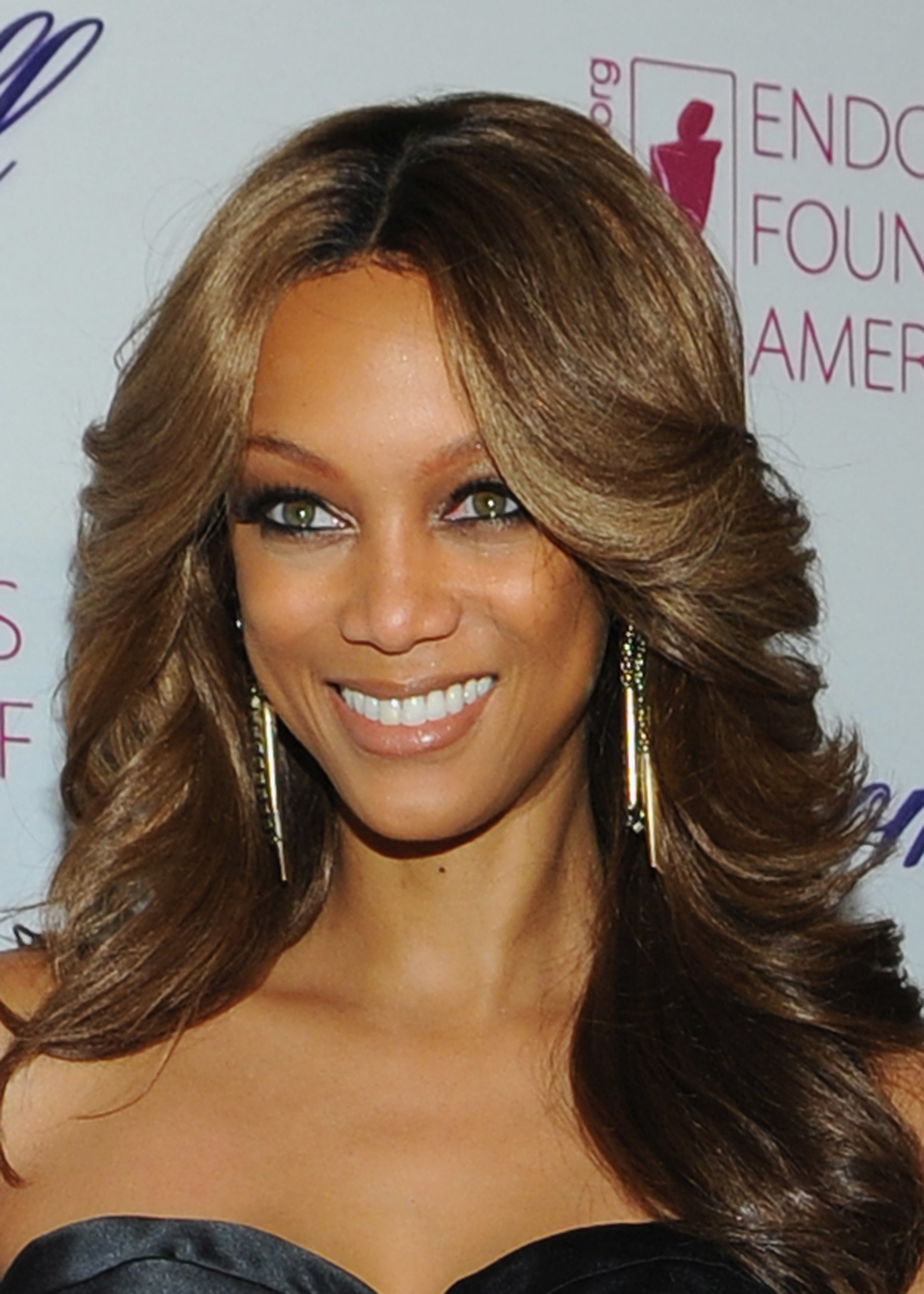 Tyra Banks smiling at an event, wearing a strapless black dress and long, gold earrings