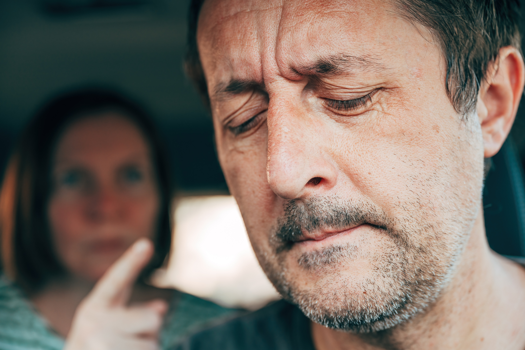 A man with a serious expression sits in the foreground, while a woman in the background points her finger at him