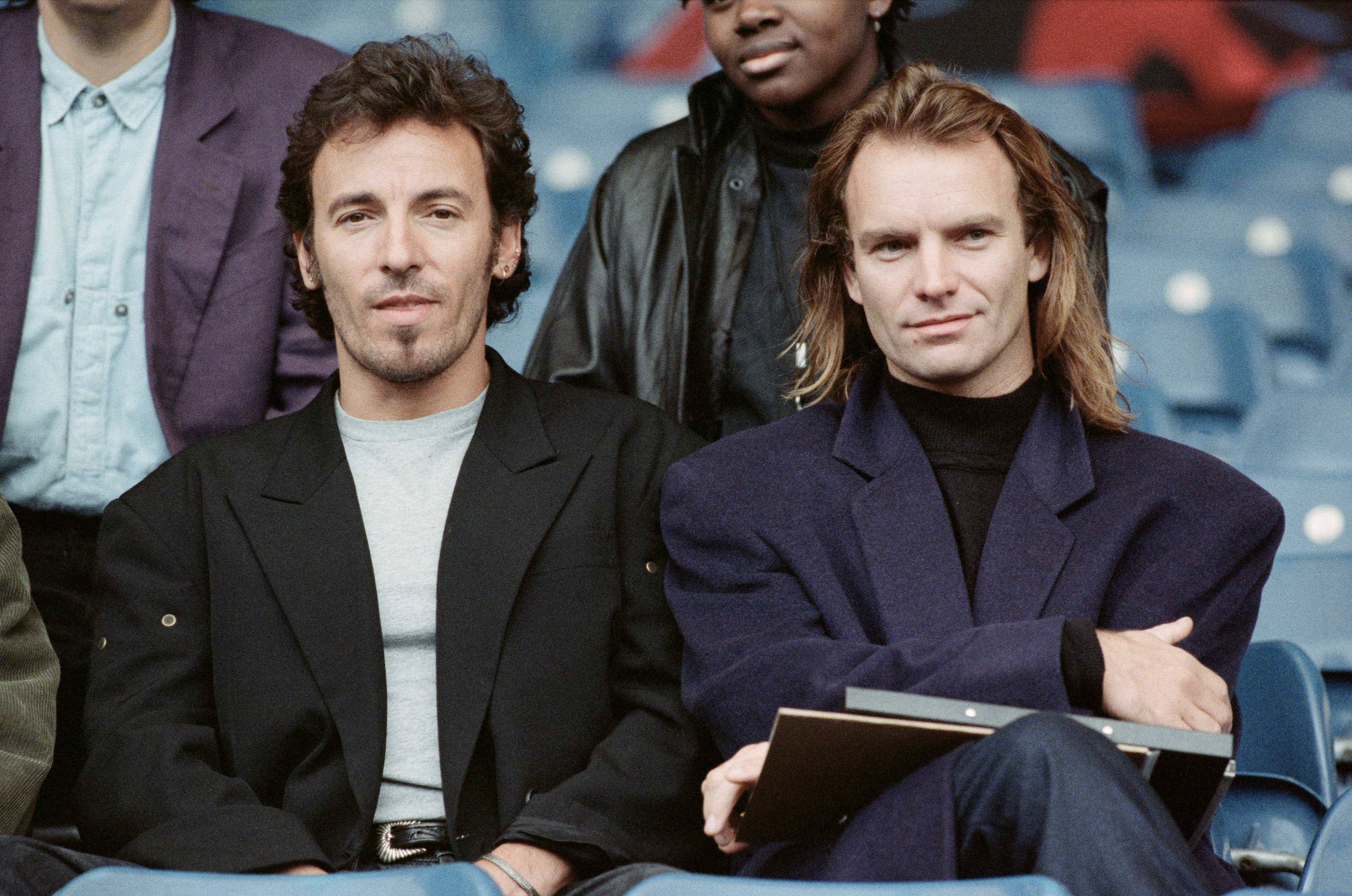 Bruce Springsteen and Sting sit next to each other, dressed in casual blazers at an outdoor event