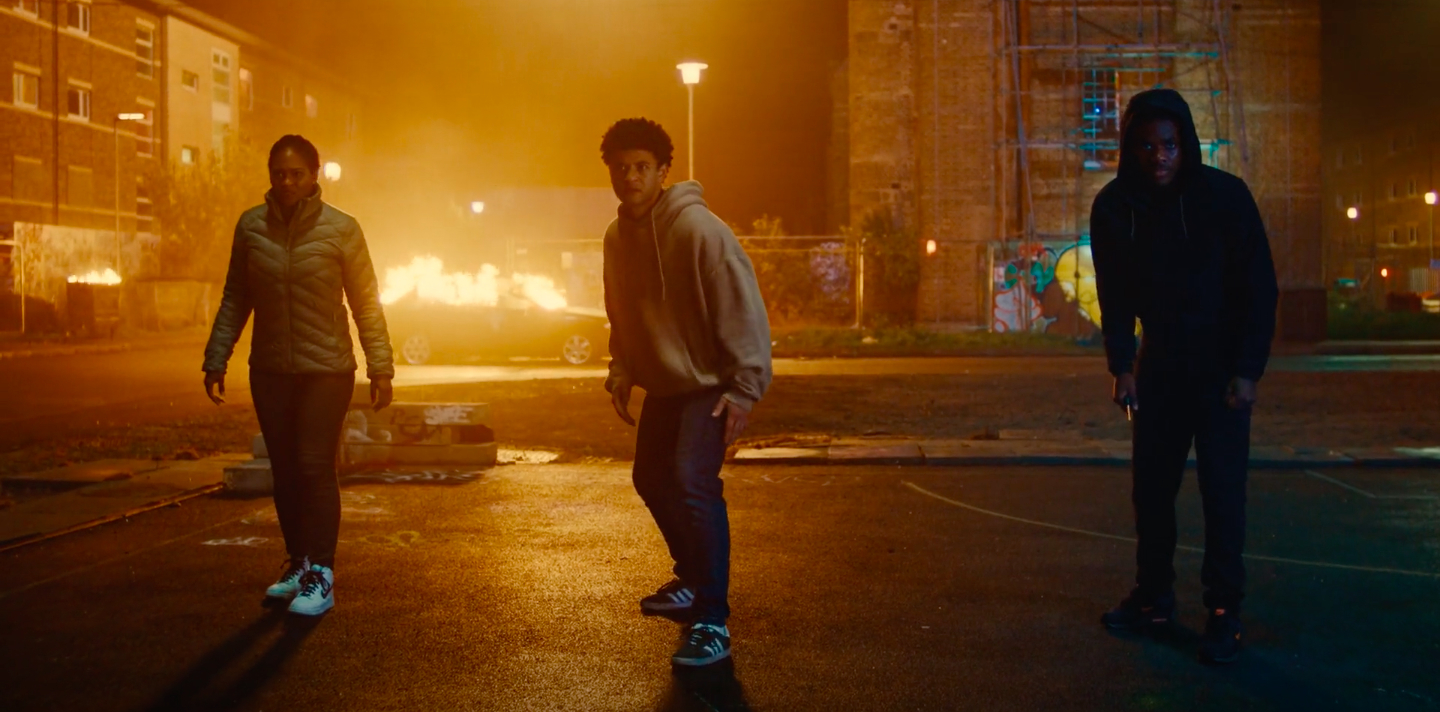 Three people in casual clothing are standing in a dimly lit urban area with a burning car in the background