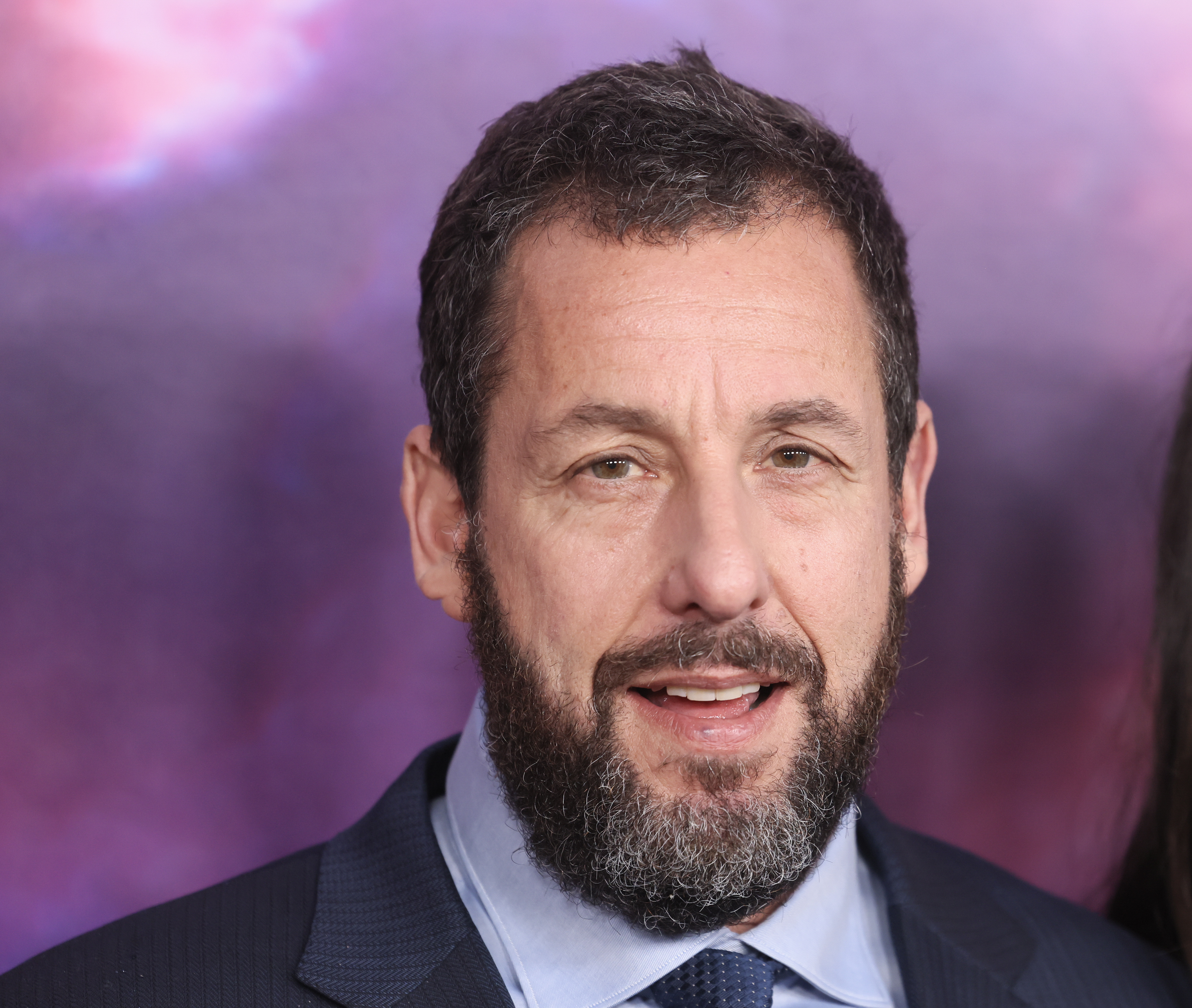 Adam Sandler at a red carpet event, wearing a suit and smiling at the camera