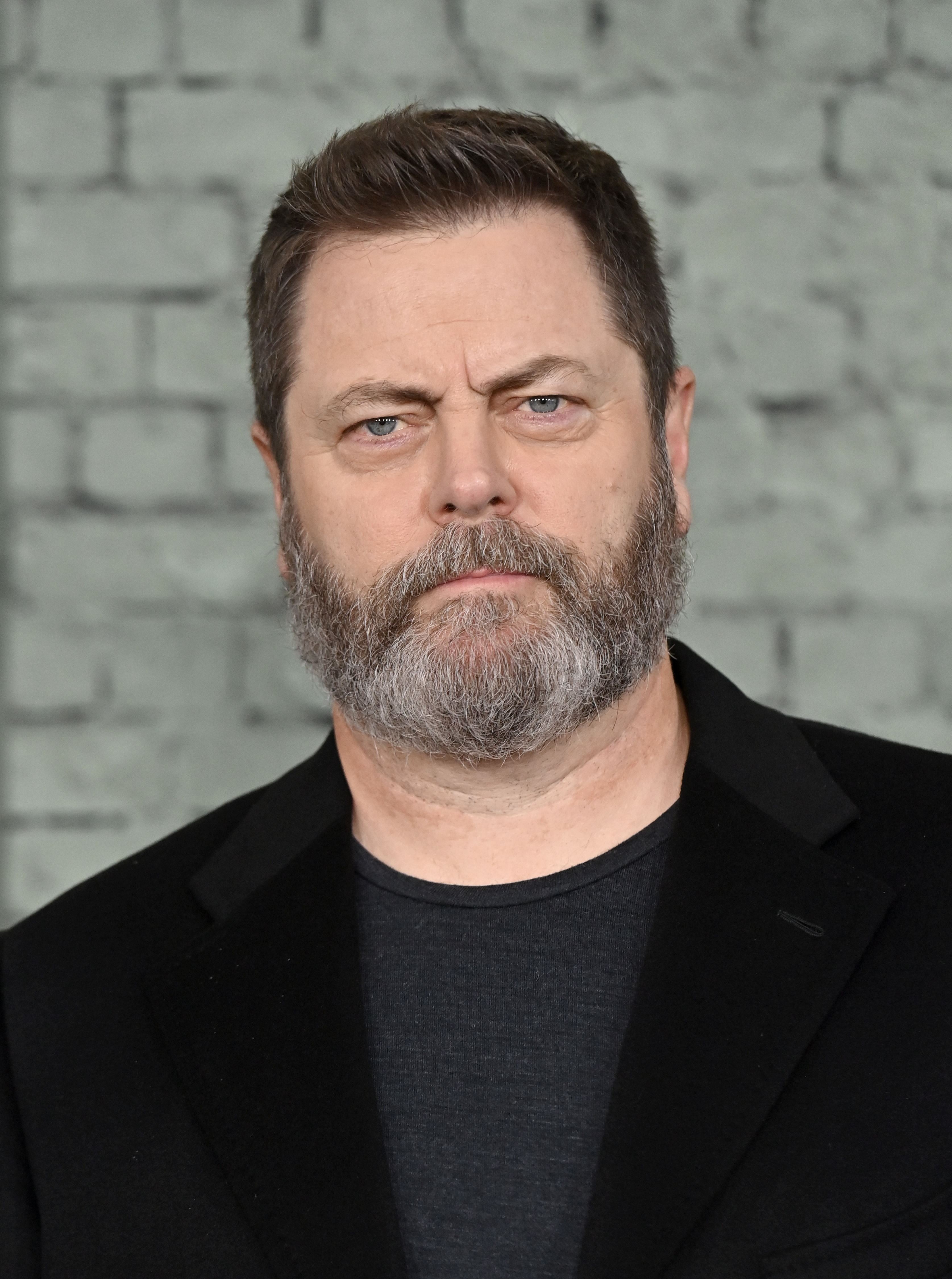 Nick Offerman poses against a brick wall backdrop, wearing a black blazer over a dark shirt