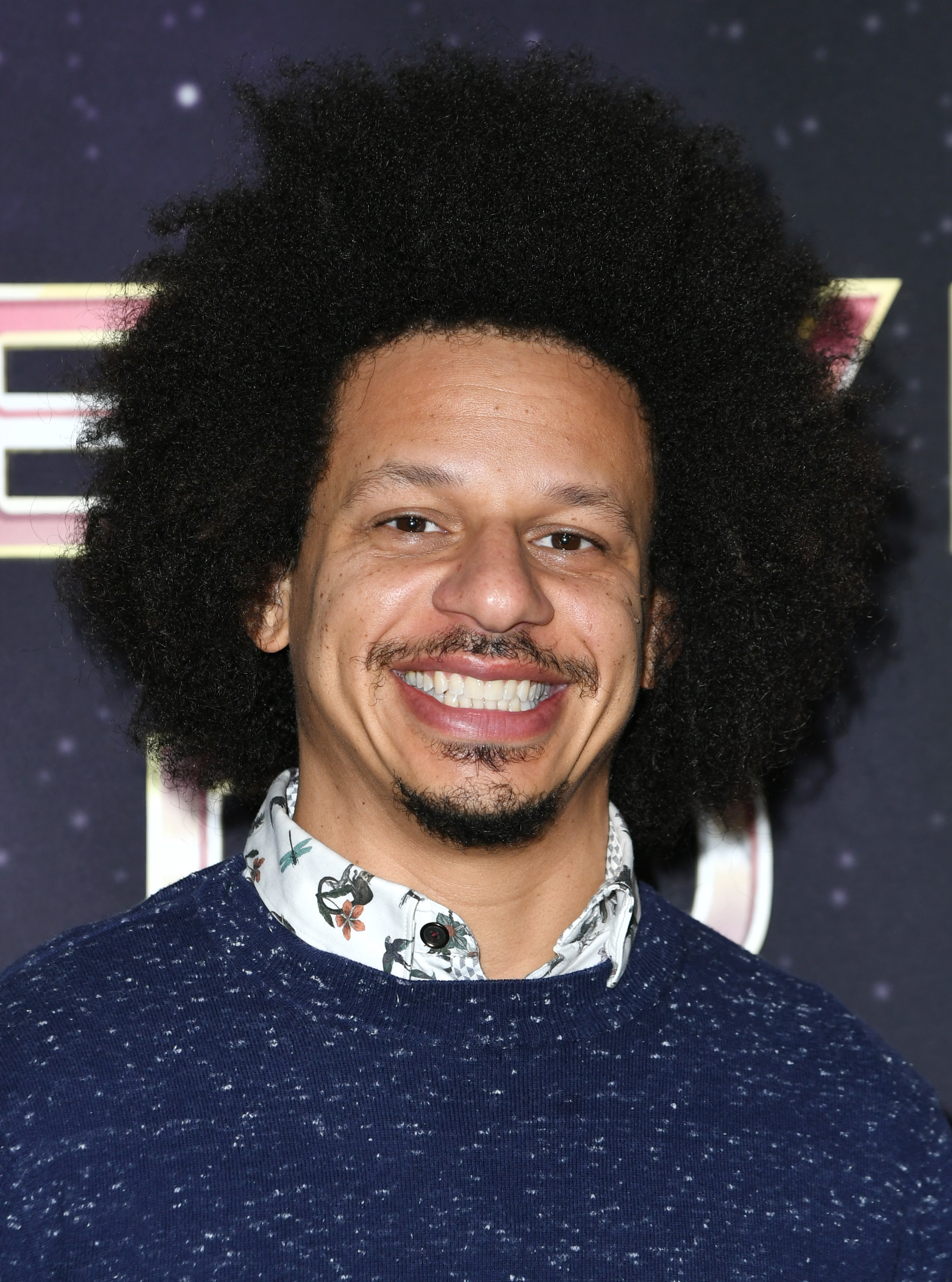 Eric Andre smiling, wearing a patterned shirt under a sweater