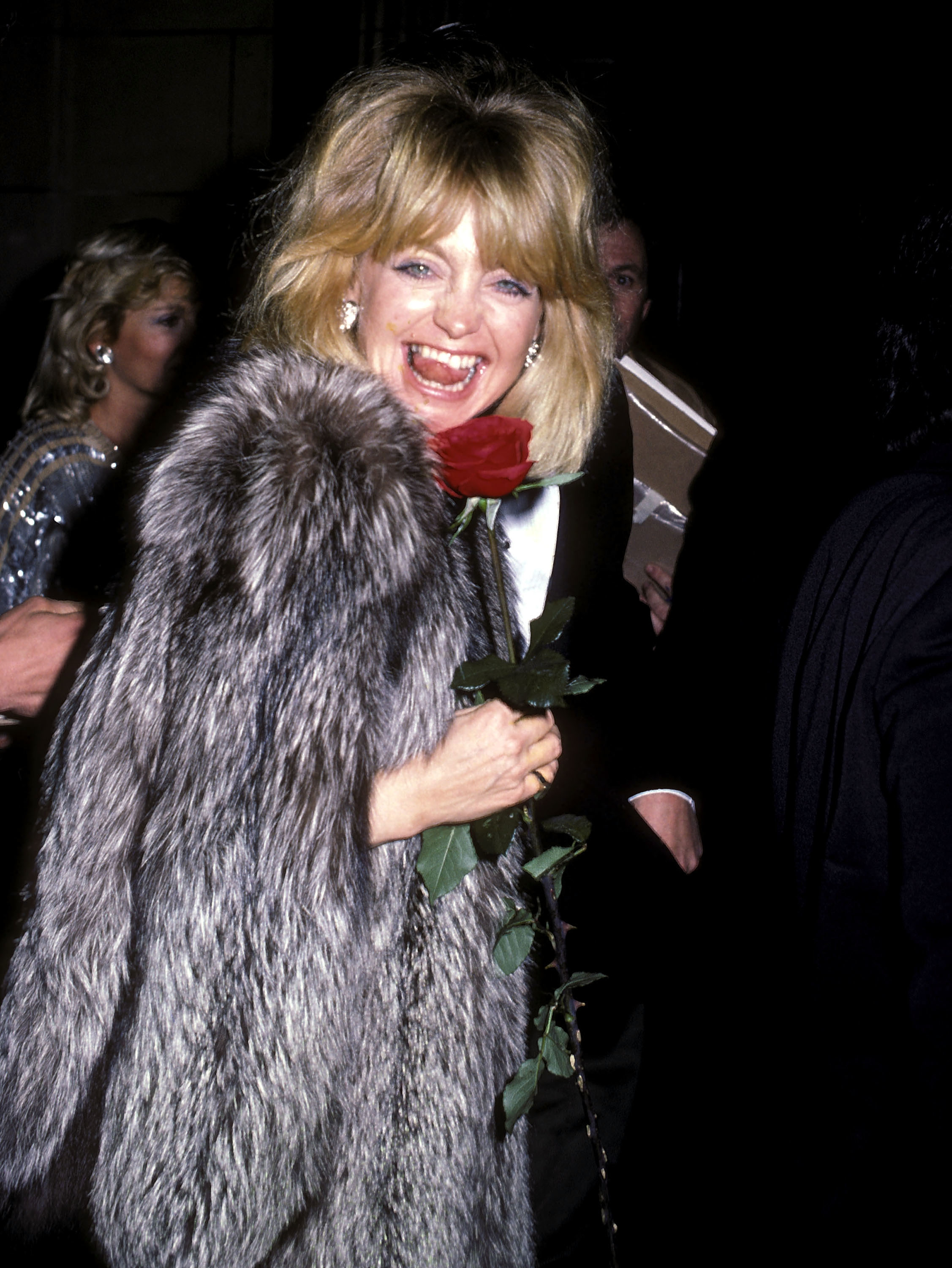 Goldie Hawn, wearing a fur coat, holds a long-stemmed rose and smiles at a formal event