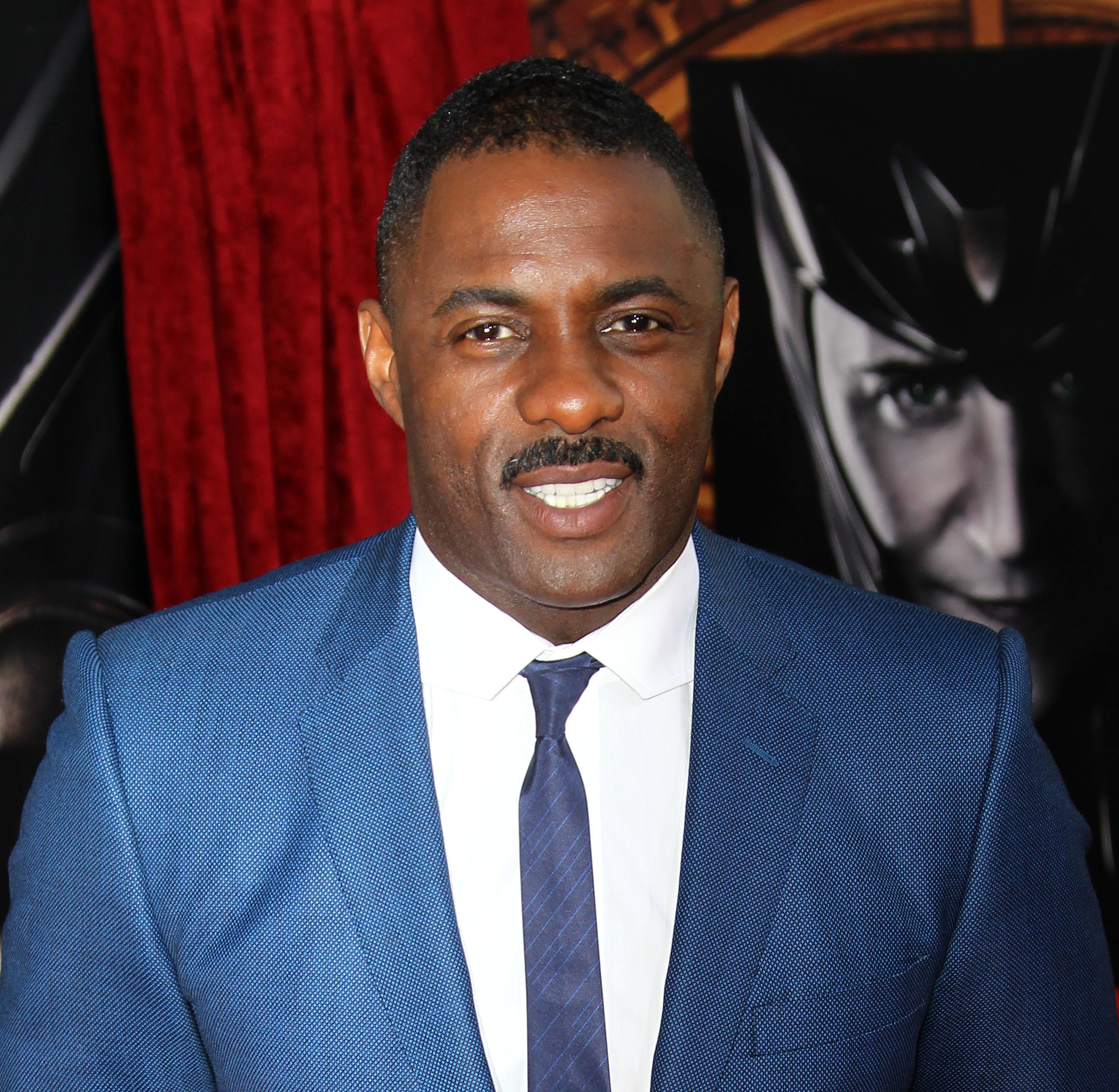 Idris Elba smiles while wearing a blue suit and tie at a red carpet event