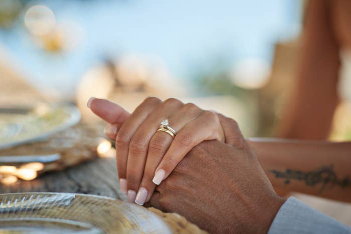 A close-up of two hands holding, showcasing an engagement ring on one hand. The hands rest on a table with plates in the background