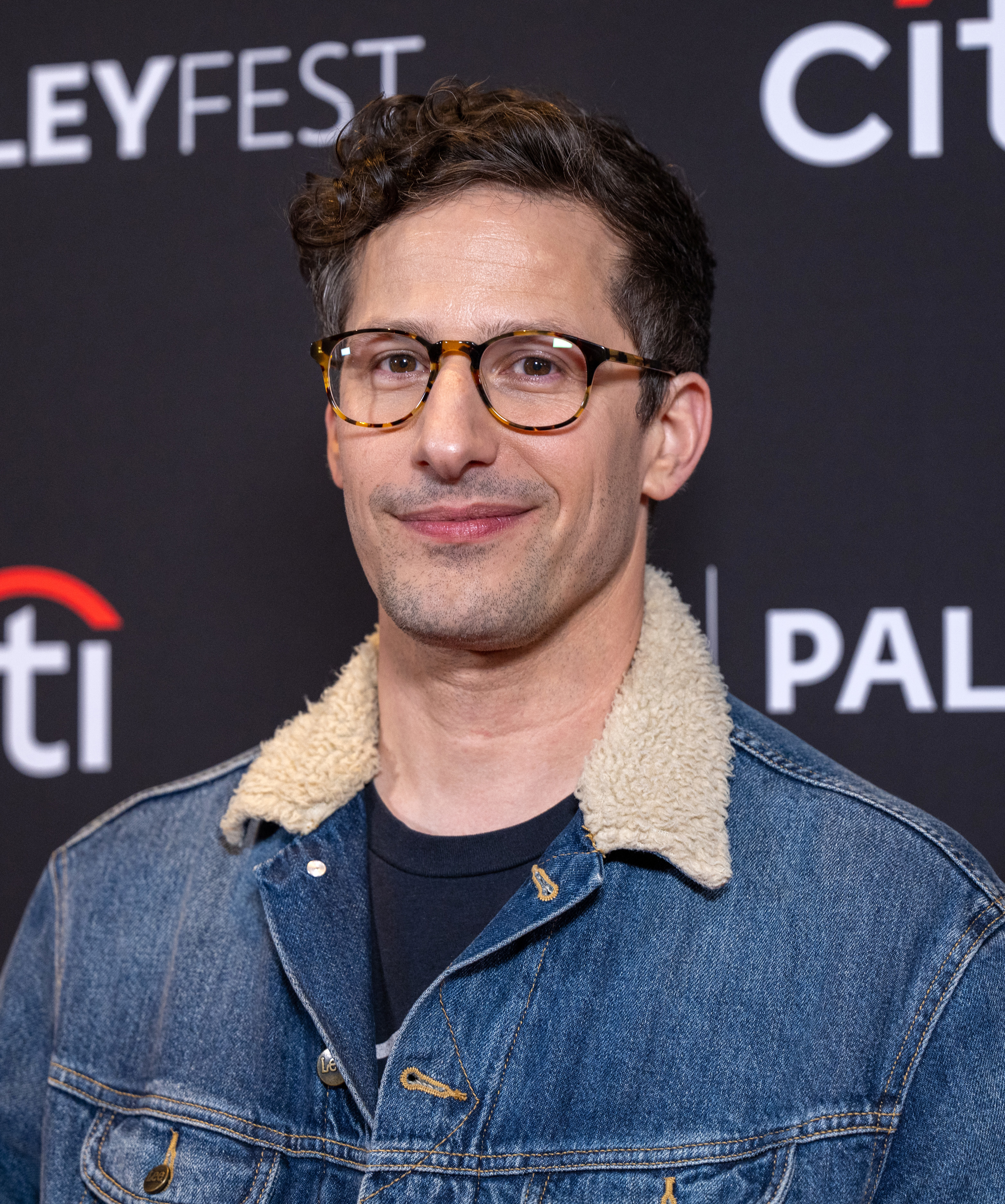 Andy Samberg at PaleyFest, wearing glasses and a denim jacket with a shearling collar, smiling for the camera