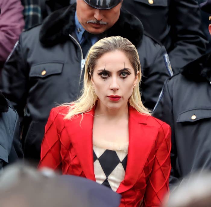 Lady Gaga in a red suit with a diamond-patterned top, flanked by police officers, likely for a film scene