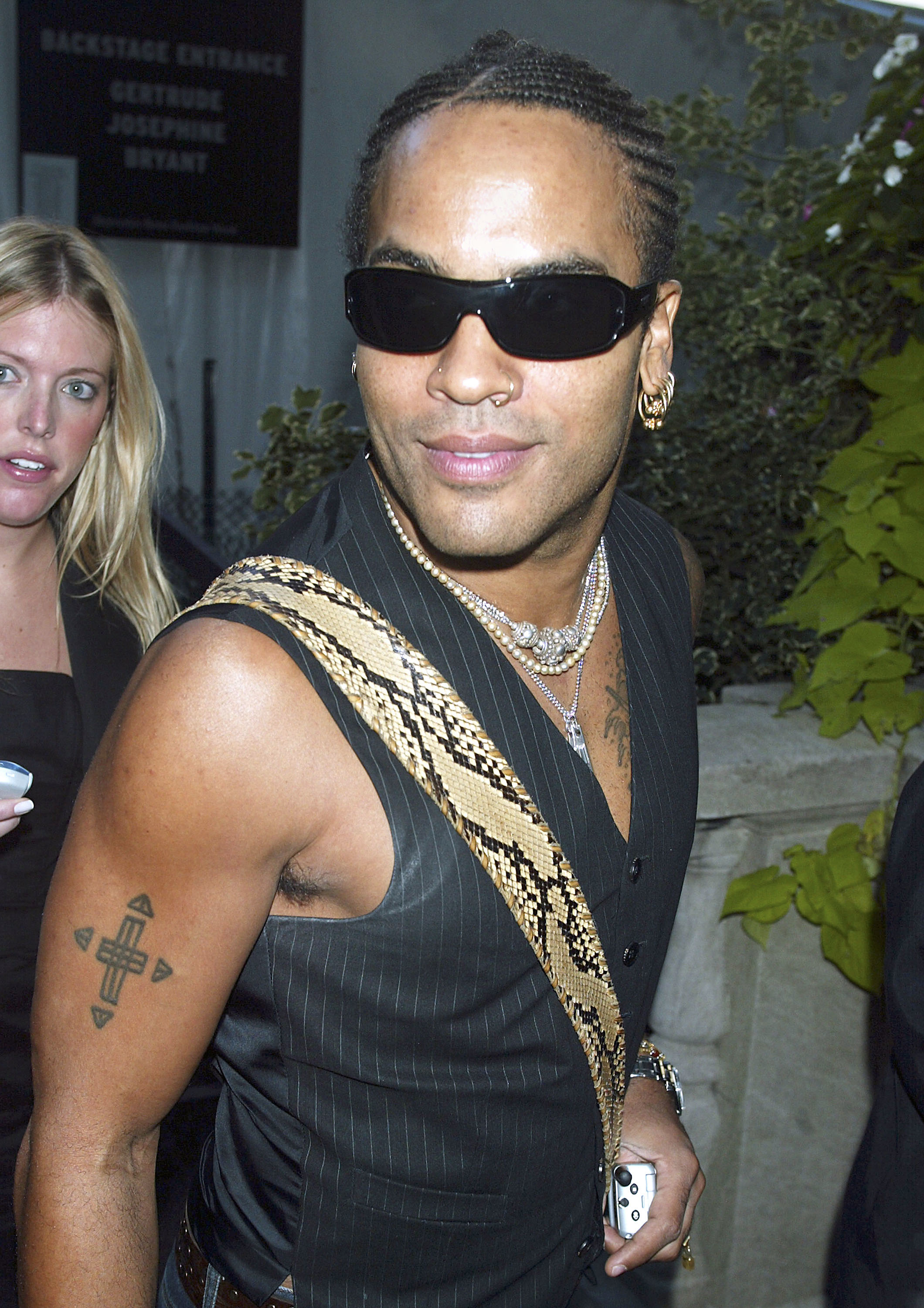 Lenny Kravitz wearing sunglasses, a black vest, and a distinctive snake-patterned strap over his shoulder. A person with blonde hair stands in the background