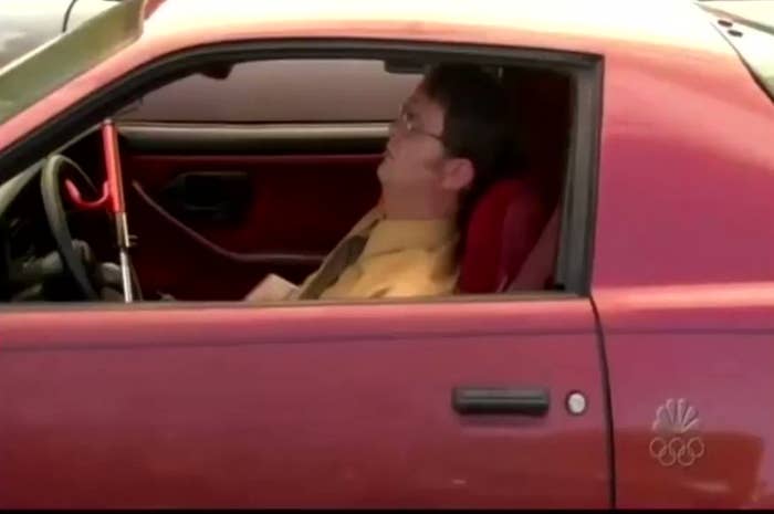 Rainn Wilson, inside a car, is sleeping with his head resting on the seat and windows rolled down. The car has a visible steering wheel lock