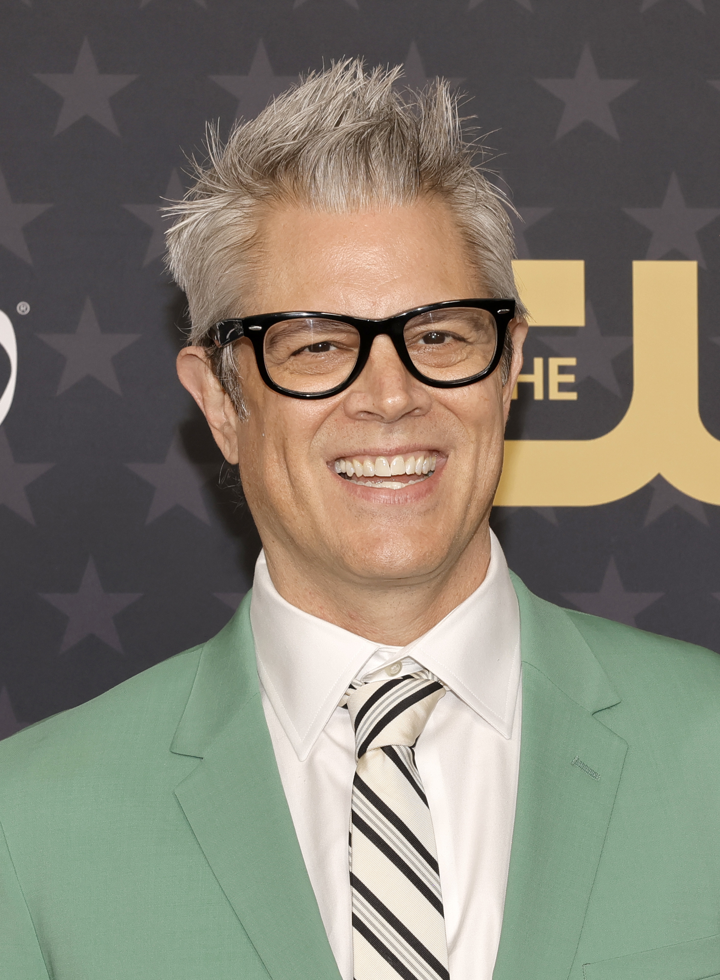 Johnny Knoxville on the red carpet, wearing a green suit, white dress shirt, and striped tie, smiling for the camera