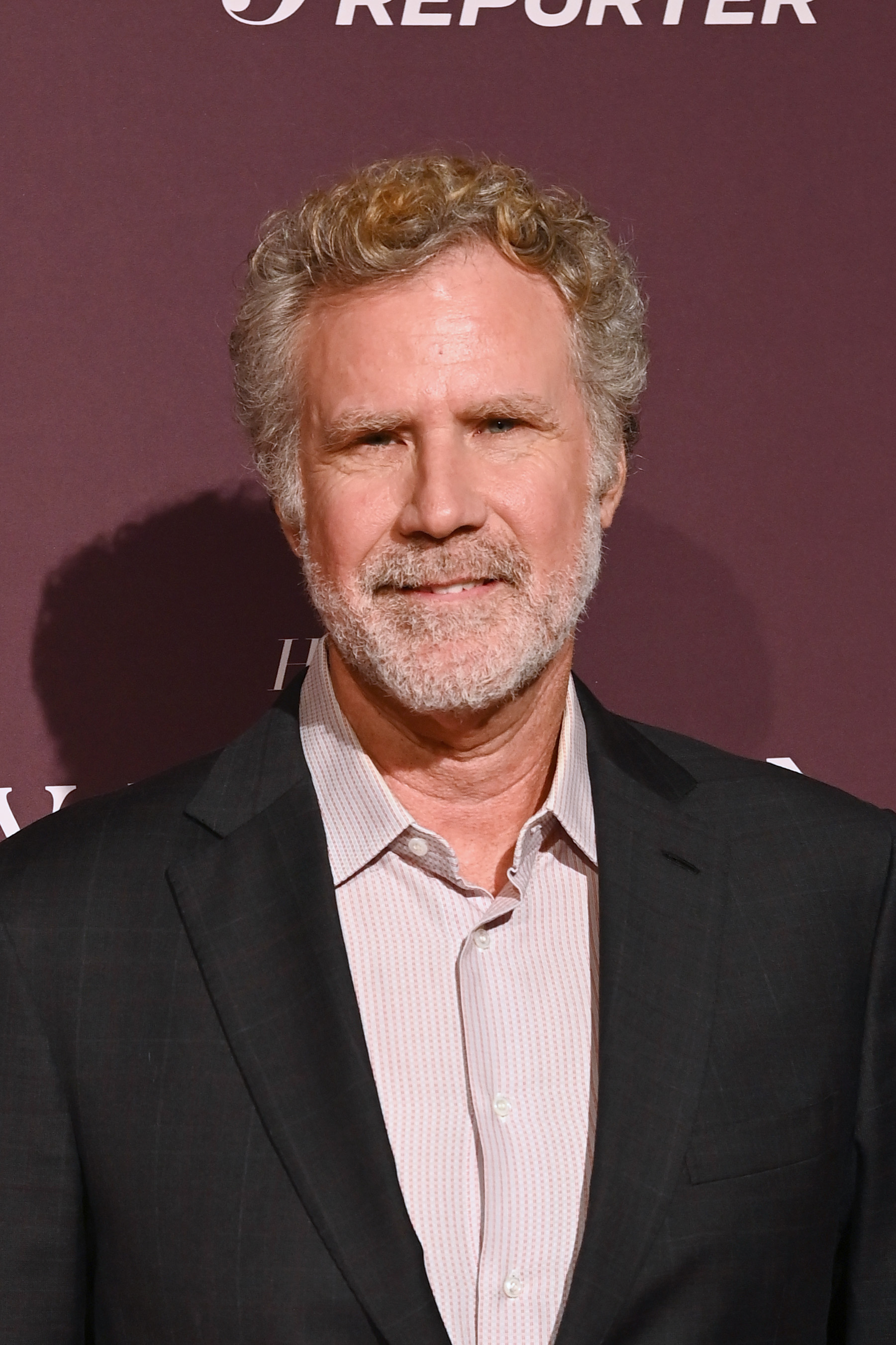 Will Ferrell at a media event, wearing a light-colored dress shirt and a dark suit jacket, standing in front of a backdrop with "Reporter" partially visible