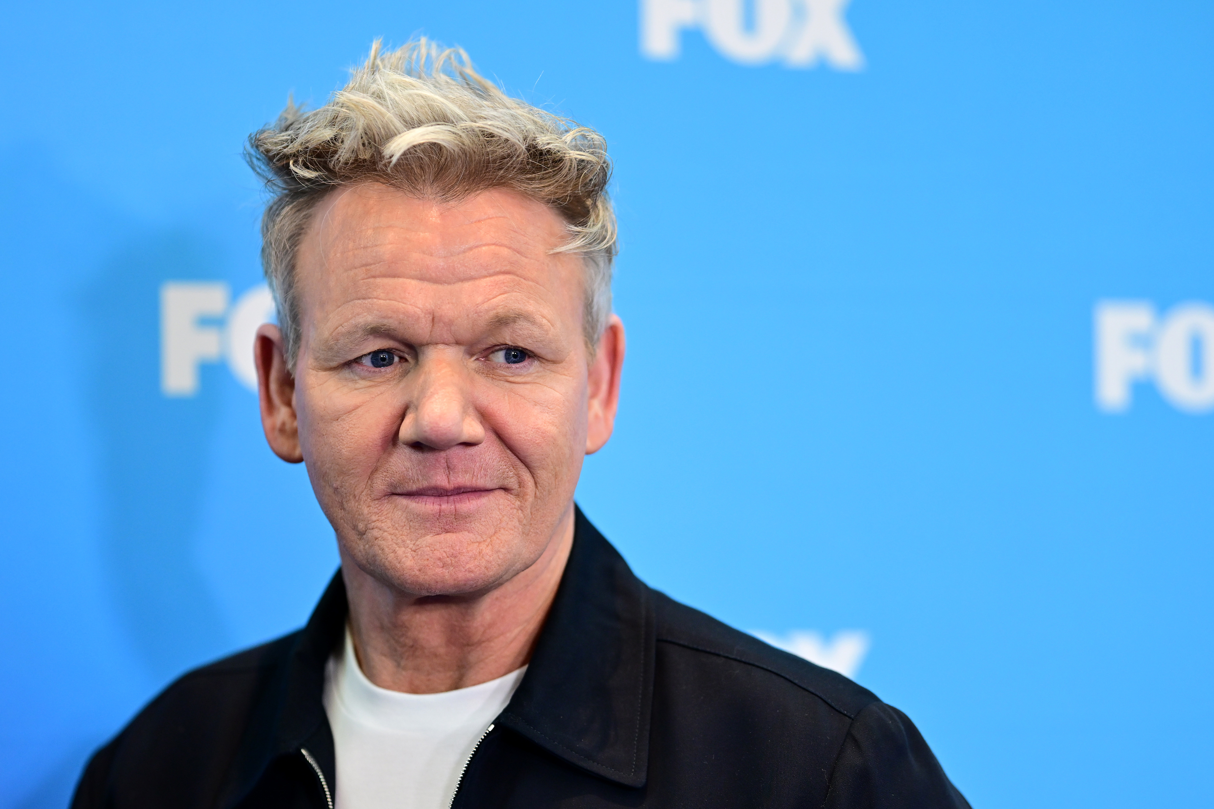 Gordon Ramsay in a black jacket over a white shirt, standing in front of a FOX backdrop