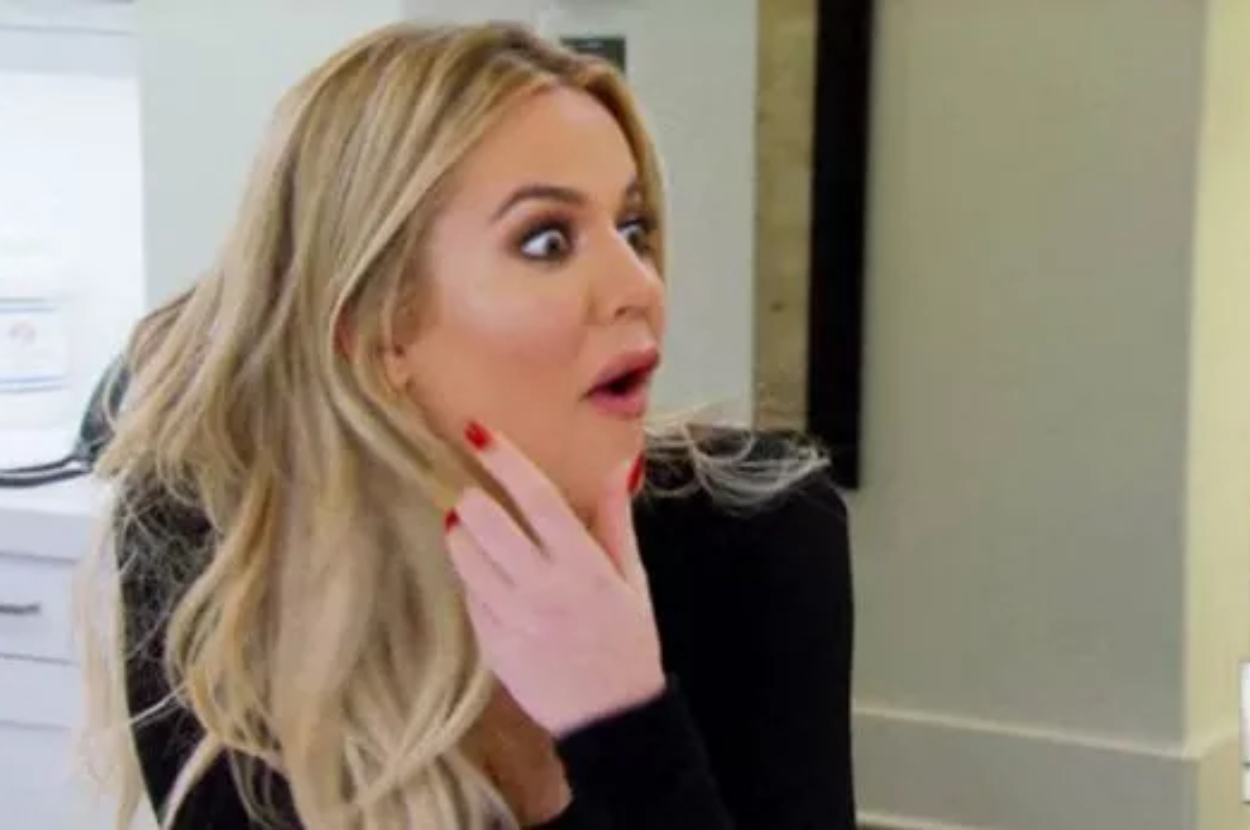 Khloe Kardashian with long hair and red nails, looking surprised with her mouth slightly open and hand touching her face