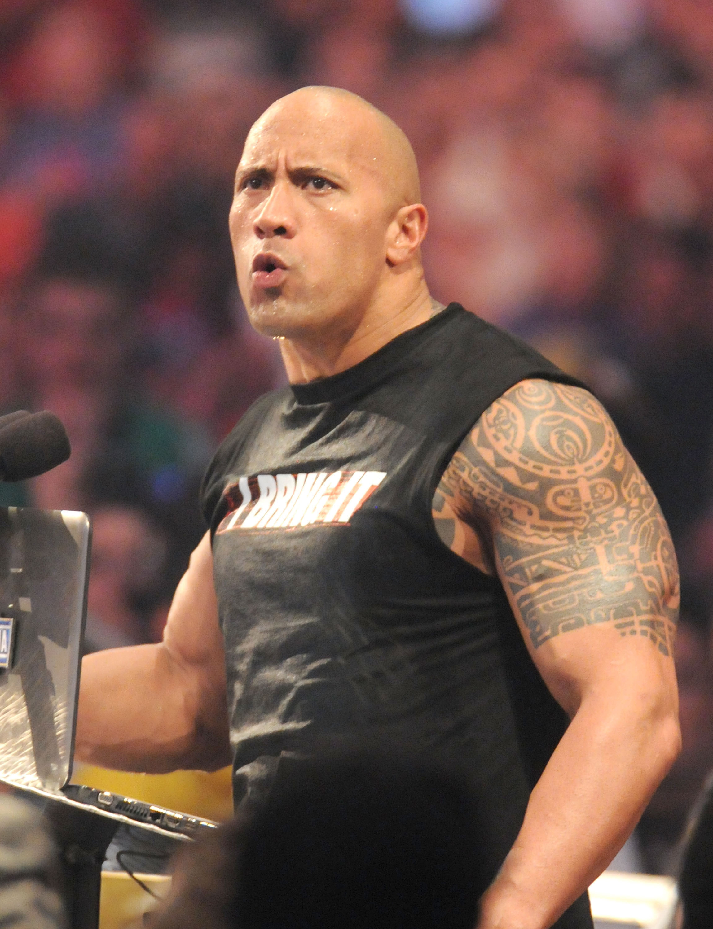 Dwayne "The Rock" Johnson wearing a sleeveless graphic tee, revealing a detailed tattoo on his left arm, appears to be speaking or performing at an event