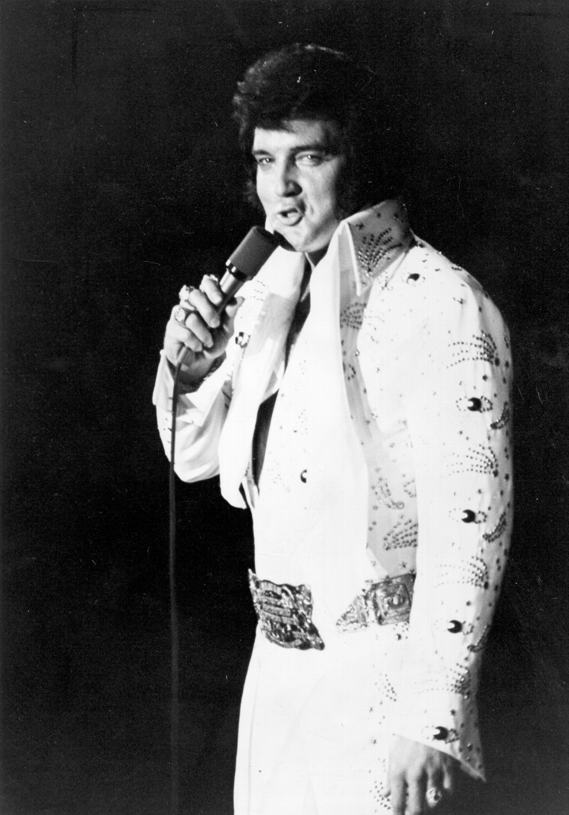 Elvis Presley singing into a microphone while wearing a white jumpsuit adorned with decorative elements and a large belt