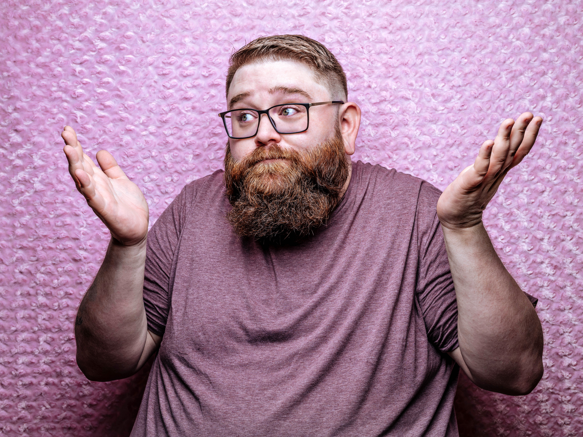 A man with a beard, wearing glasses and a t-shirt, stands against a textured backdrop with a puzzled expression, shrugging his shoulders. Names not known