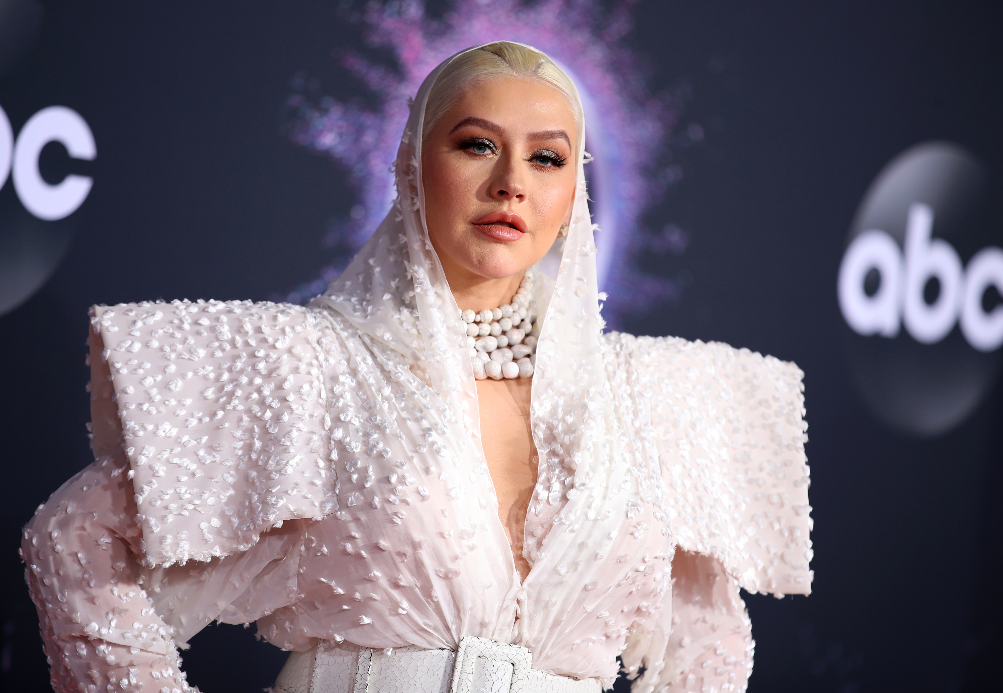 Christina Aguilera on the red carpet, wearing a beaded, textured outfit with a headscarf and pearl necklace