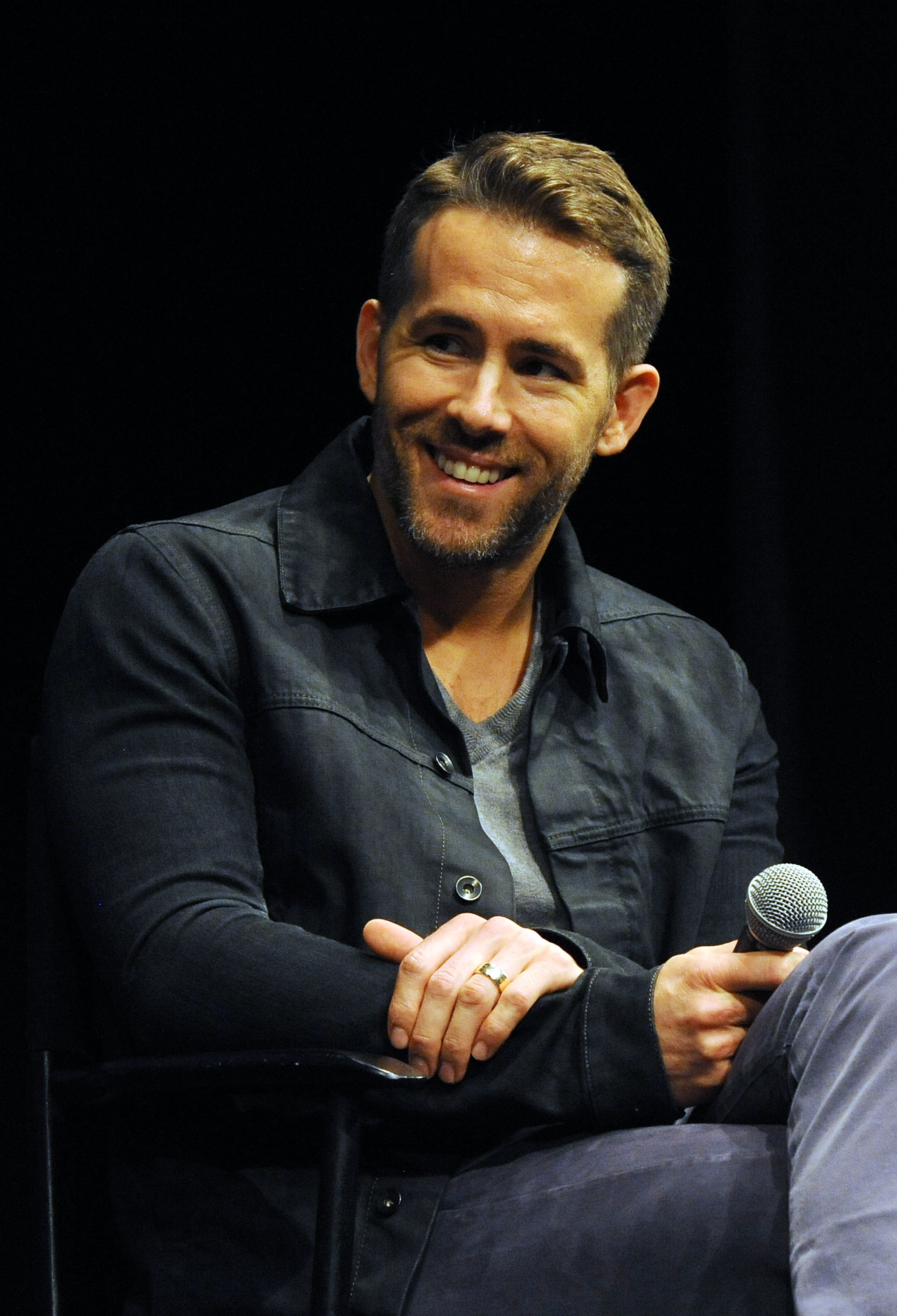 Ryan Reynolds sits smiling in a casual jacket, holding a microphone during an event